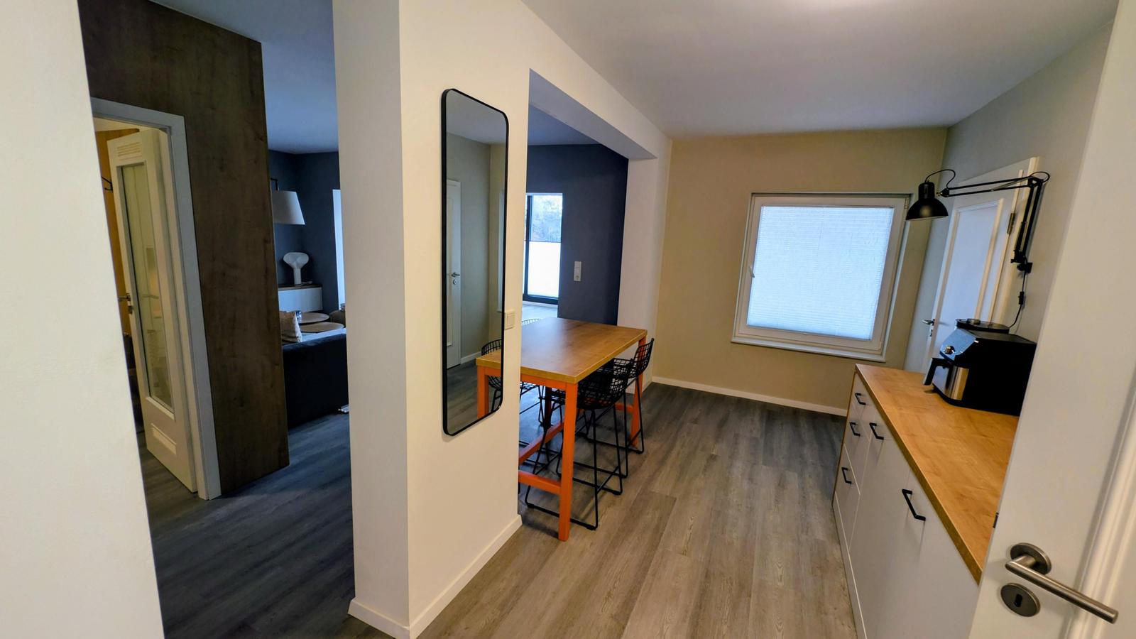 Küche mit Holzplatte und Stuhl. Tisch mit Stühlen. Spiegel an der Wand. Tür zum Schlafzimmer.