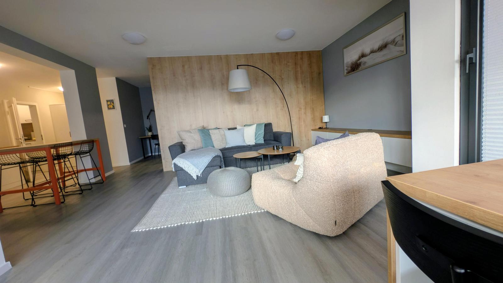 Wohnzimmer mit Sofa, Sessel und Holzwand. Küche und Essbereich sind sichtbar.