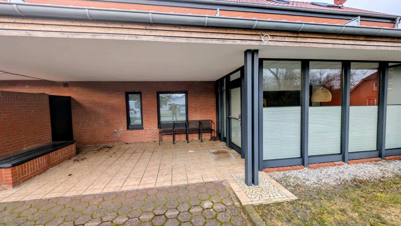 Terrasse mit Sitzbank, Glasfront und Backsteinmauer. Eingang mit Rollladen.