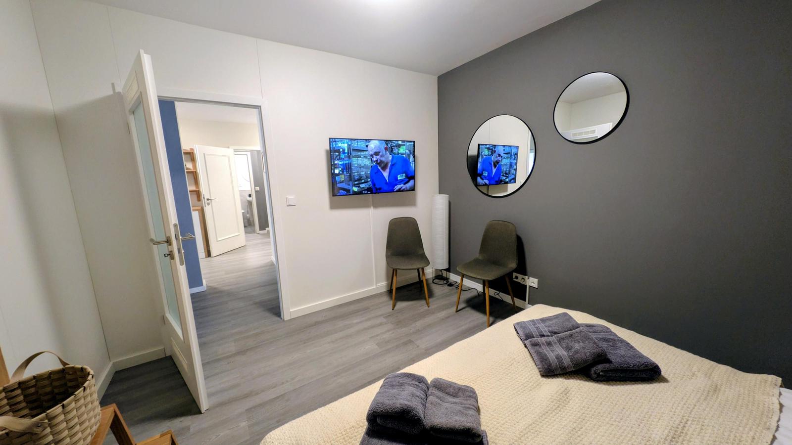 Schlafzimmer mit Bett, Fernseher und zwei Stühlen. Zwei runde Spiegel an der Wand.