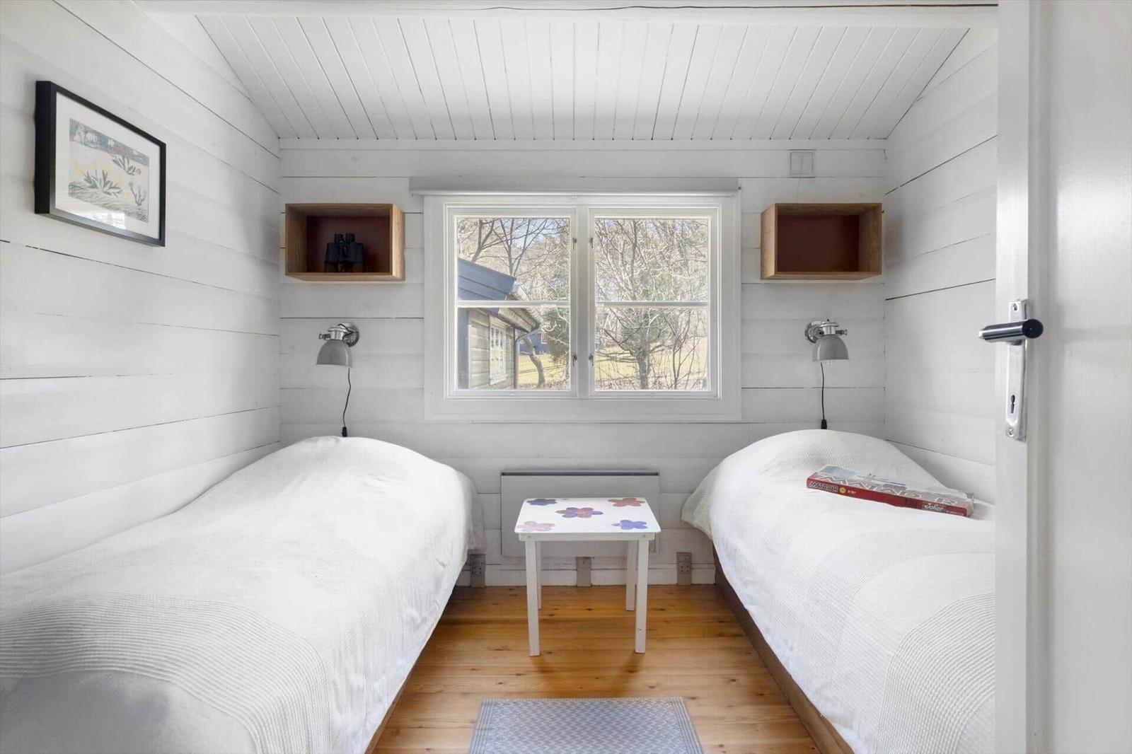 Two beds, window, table, and wall shelves in a room with white wood.