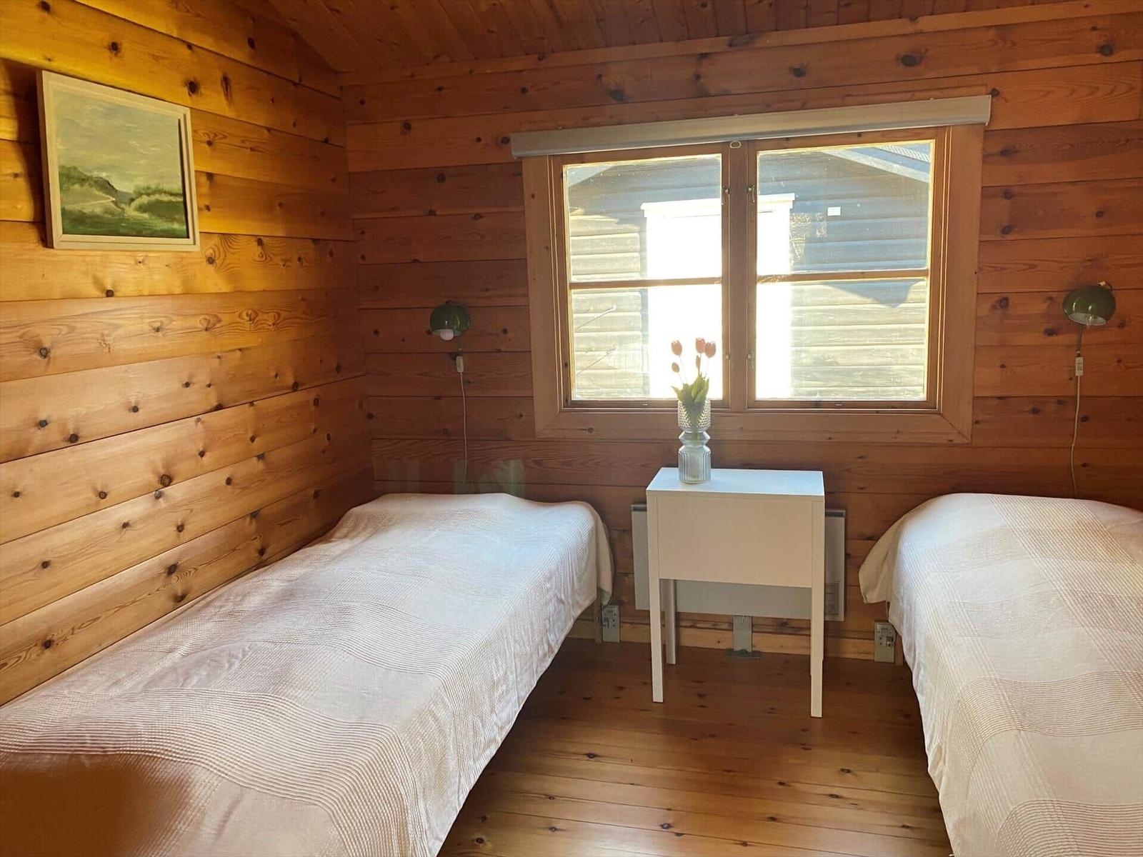 Two beds, a nightstand with flowers, wooden walls, and window.