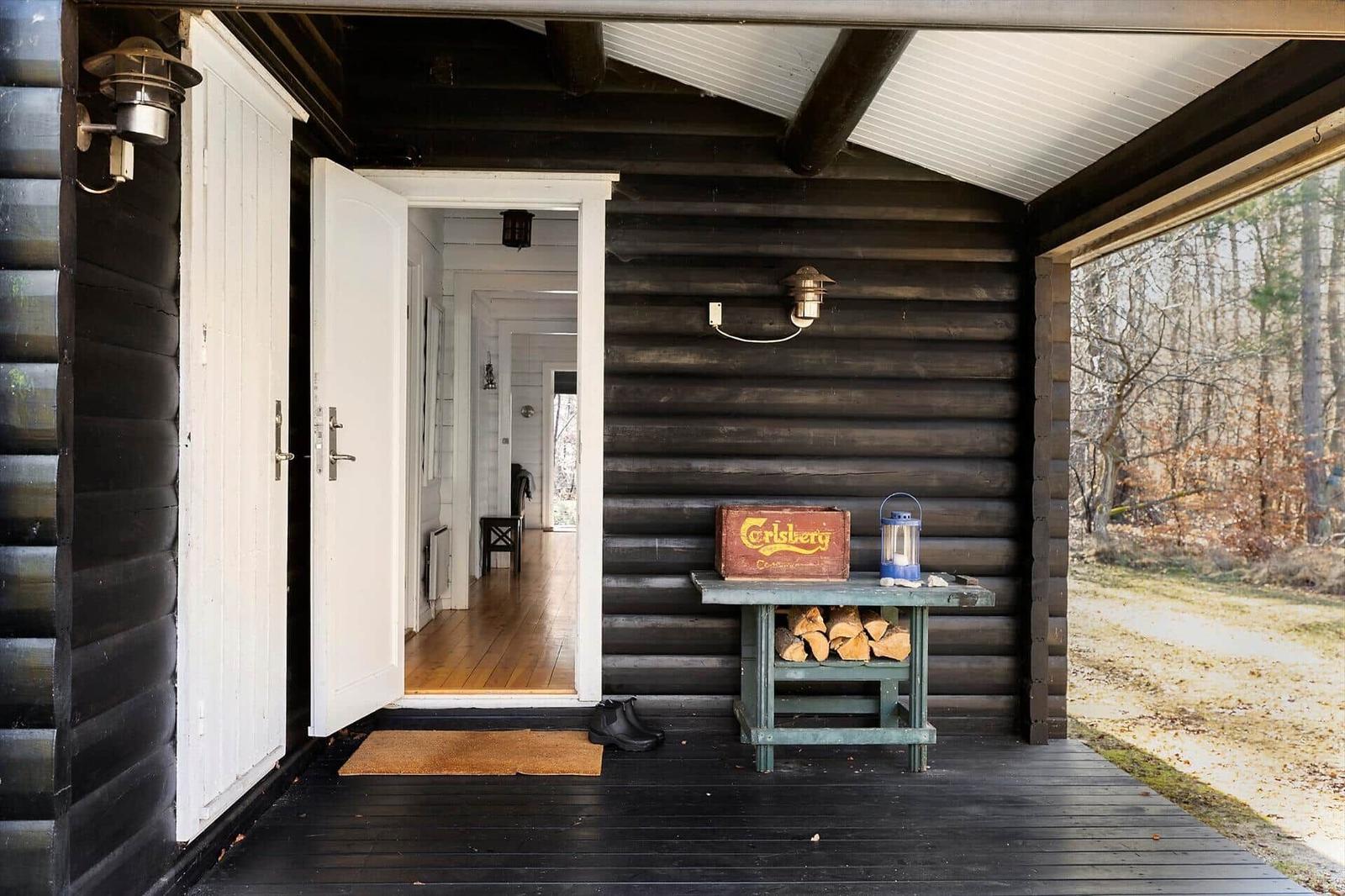 Entrance of a log cabin with open door and stacked firewood. Forest landscape in the background.