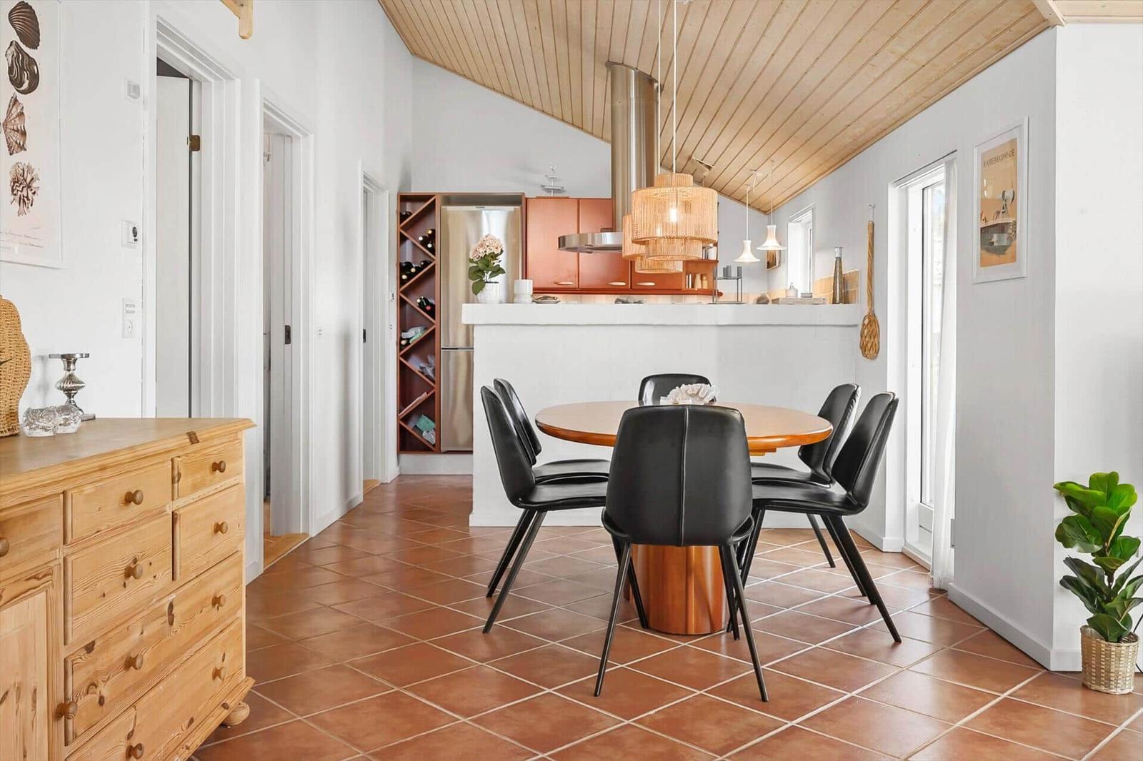 Dining area with round table and black chairs. Kitchen area with wooden ceiling and red tiles.