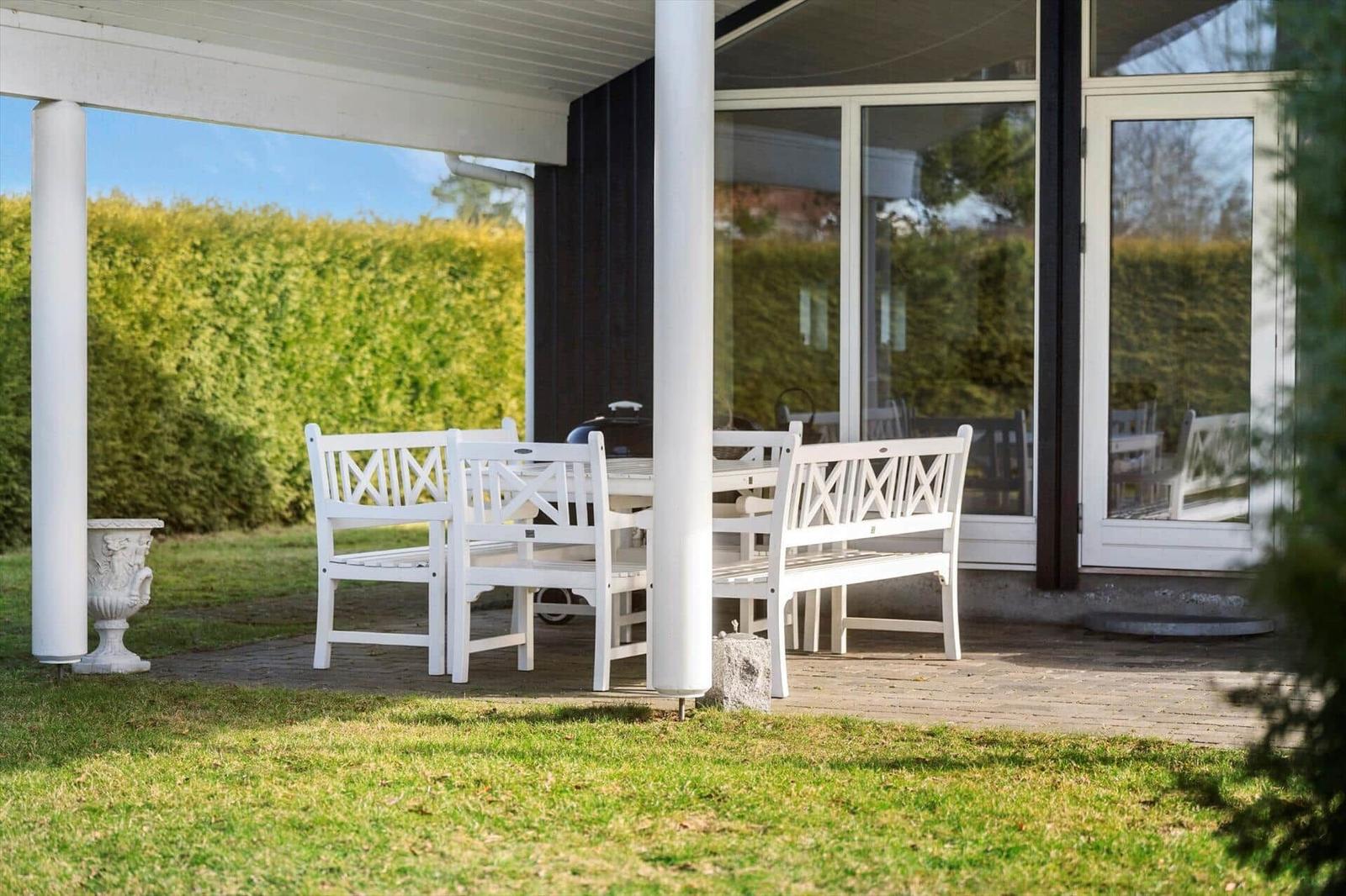 White outdoor area with table and chairs under cover. Background: green hedge area.
