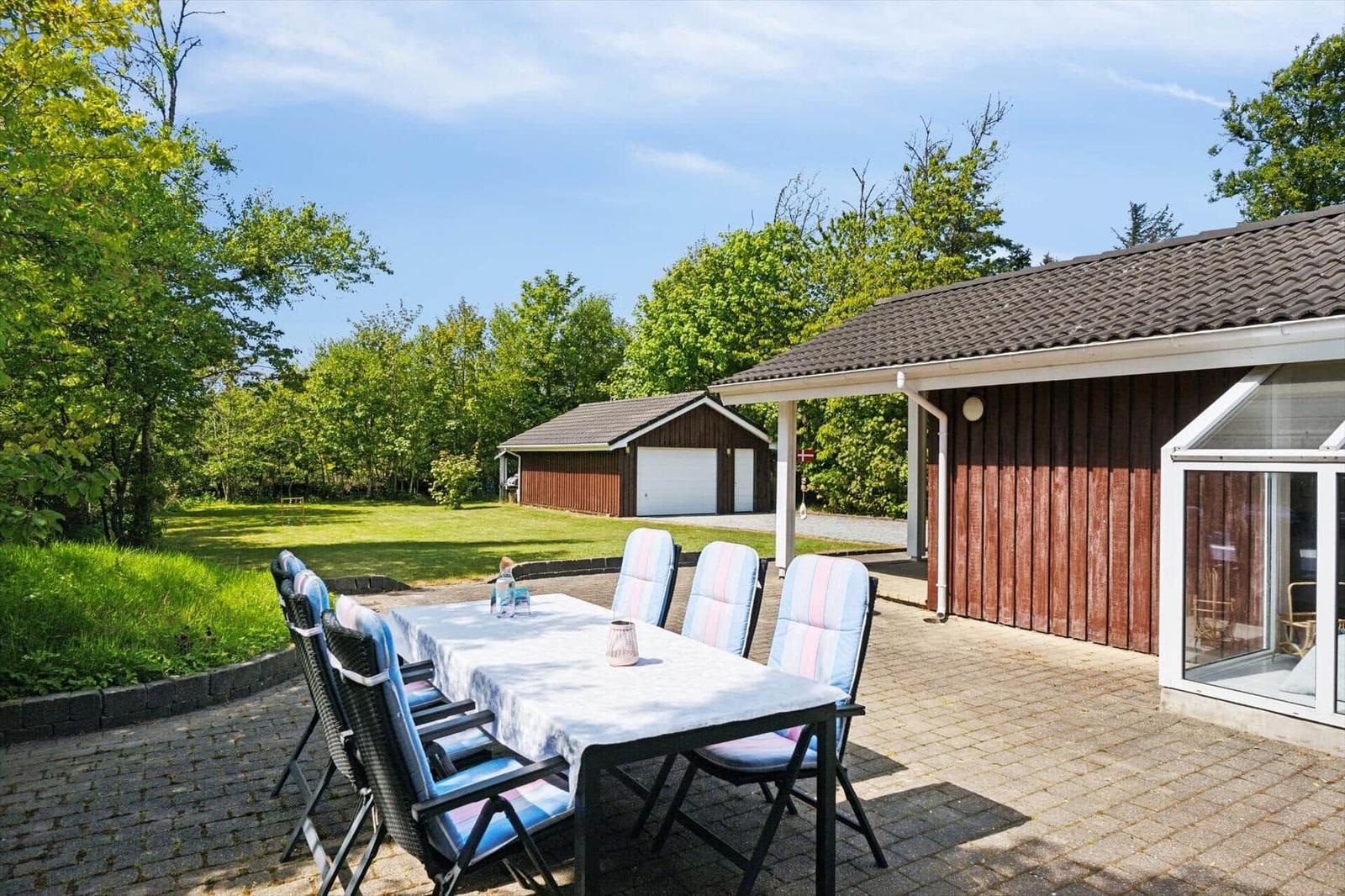Terrace with table and chairs beside a wooden house. Lawn and trees in the background.