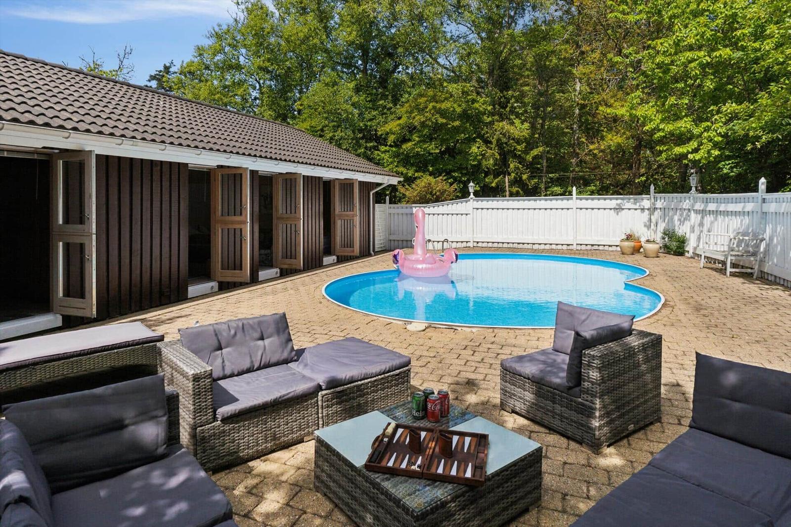 Pool with pink float, patio with seating and wooden house in background.