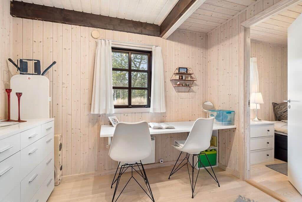 Wohnraum mit Schreibtisch, zwei Stühlen und Fenster mit weißen Vorhängen.
