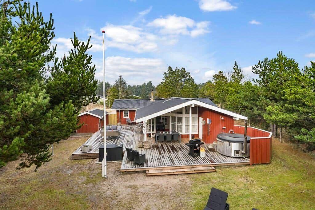 Rotes Ferienhaus mit Terrasse, Whirlpool und Grill unter Bäumen.