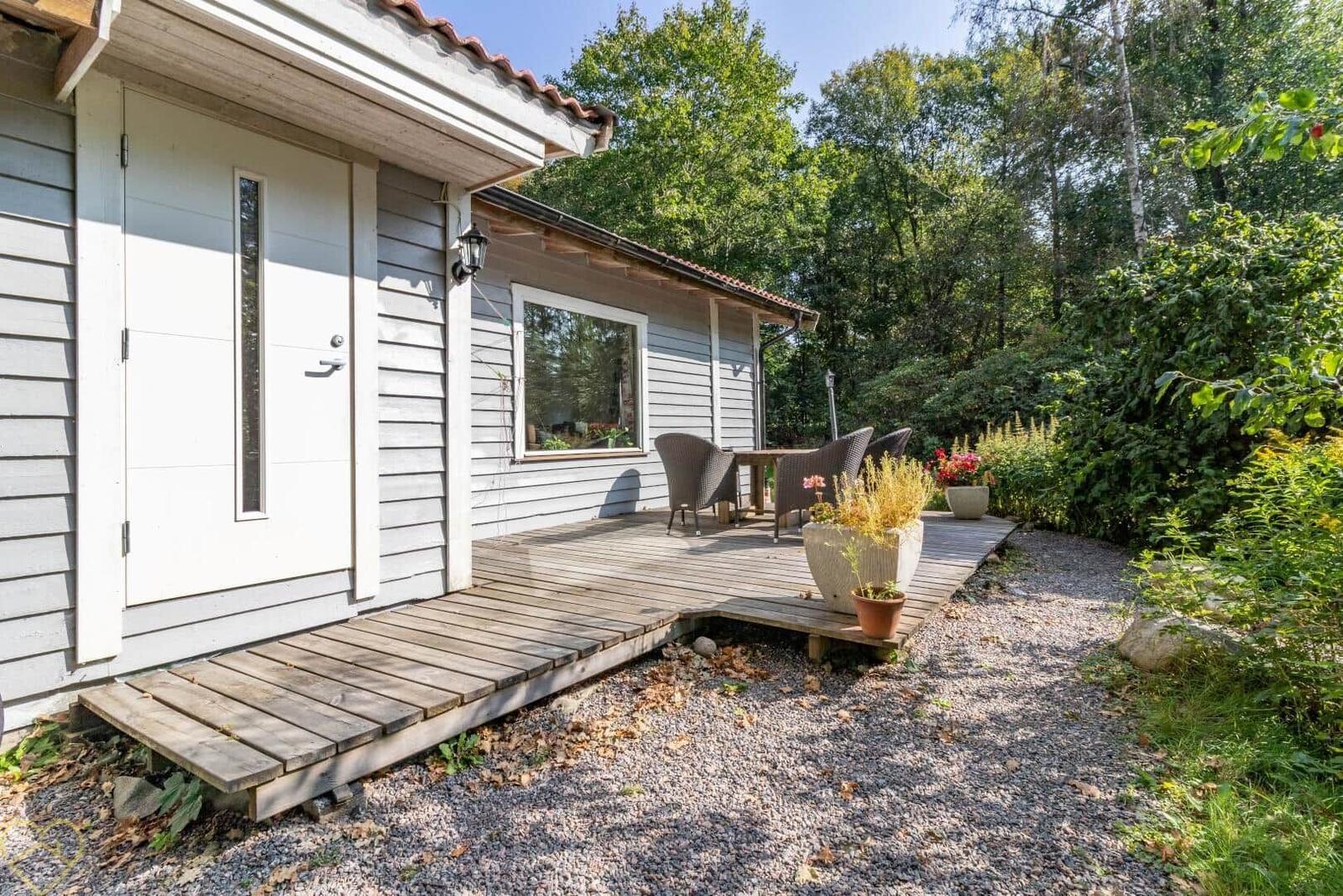 Haus mit Terrasse, Tisch und Stühlen, umgeben von Bäumen und Pflanzen.