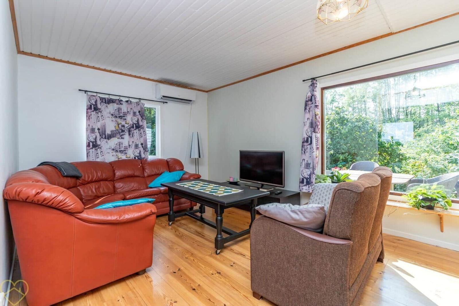 Wohnzimmer mit rotem Sofa, braunem Sessel und Holzboden. Fenster mit Blick auf Garten.