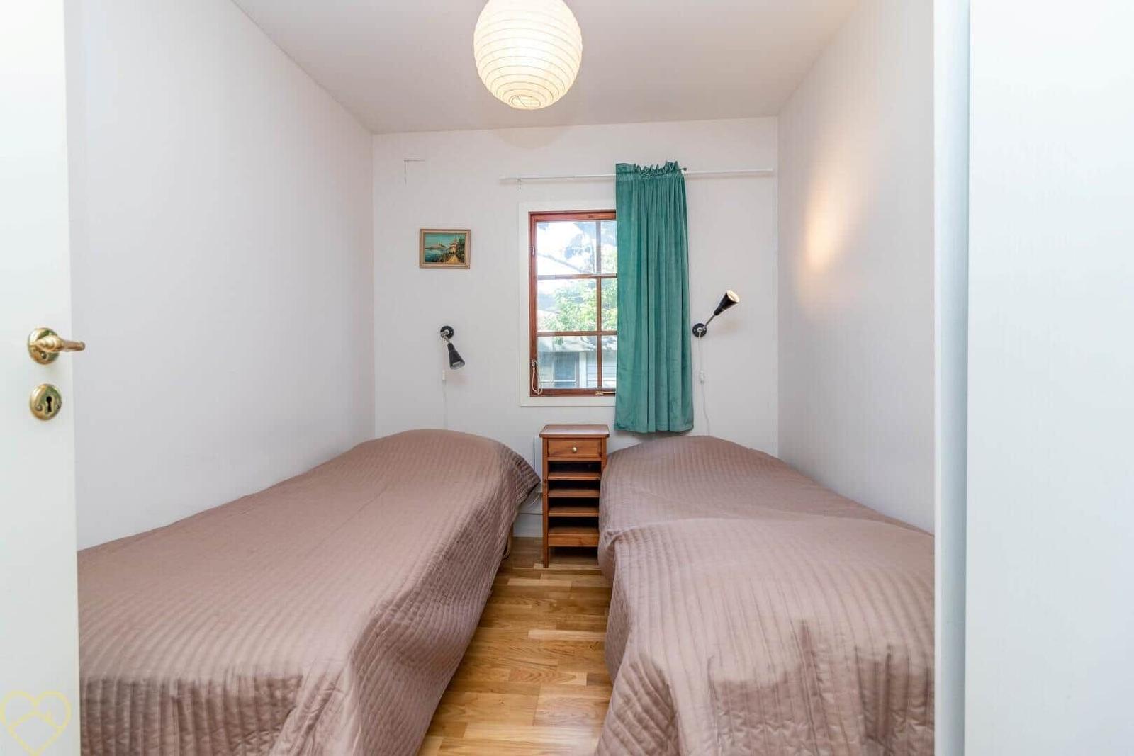 Zwei Betten mit braunen Bettdecken, Holzfußboden und Fenster mit grüner Gardine.