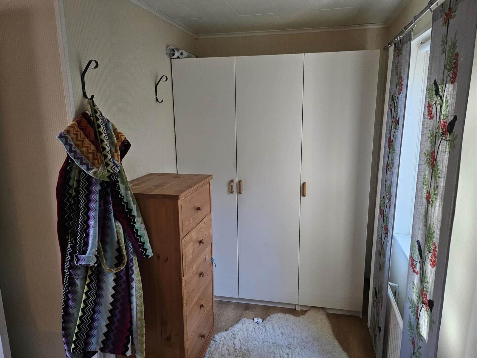 Schlafzimmer mit Kleiderschrank, Holzkommode und Fenster mit Vorhängen.