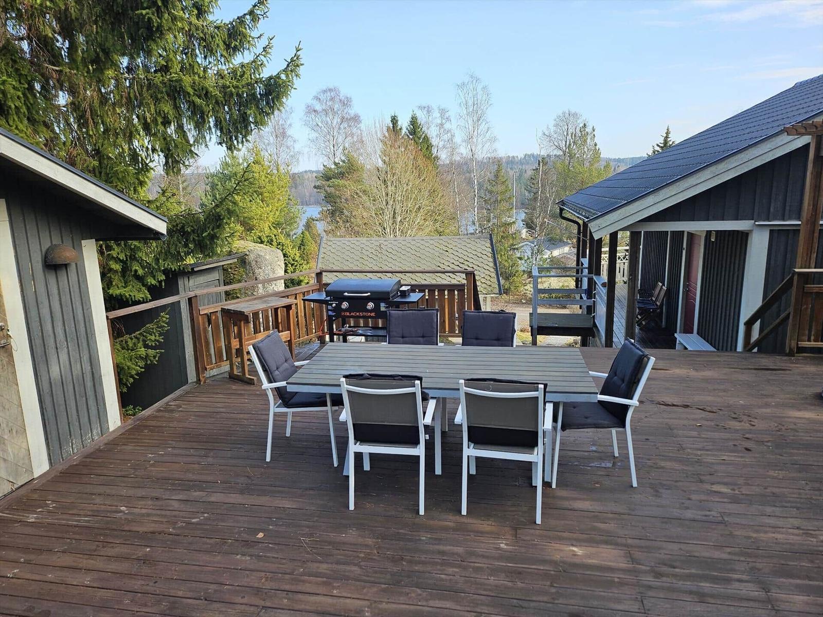 Holzterrasse mit Tisch, Stühlen und Grill. Blick auf Wald und Wasser.