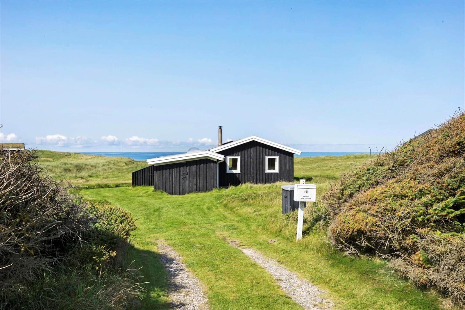 Schwarze Hütte mit weißen Fenstern, umgeben von Gras und Büschen, Blick auf das Meer.