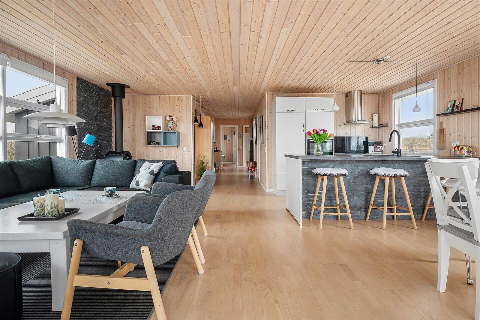 Living and kitchen area with wooden floor, ceiling, and walls. It has a sofa, chairs, and a kitchen island.