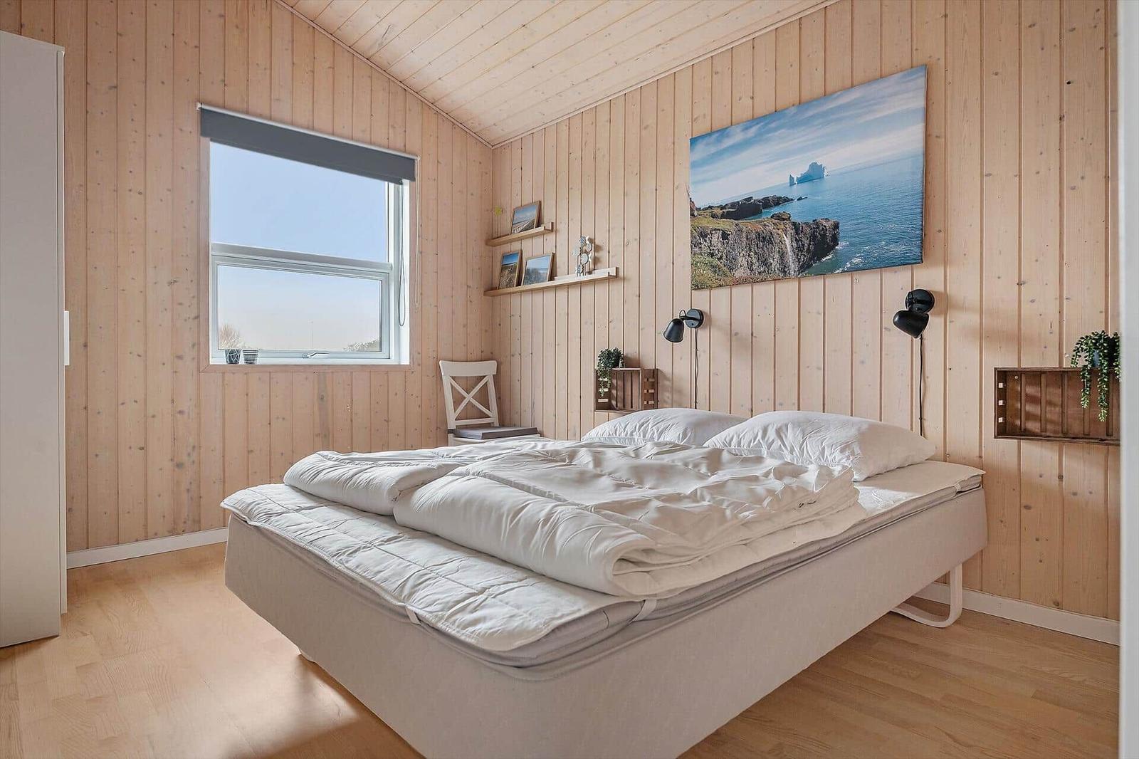 Bedroom with wooden walls, bed, and window. Wall art shows coastal landscape.