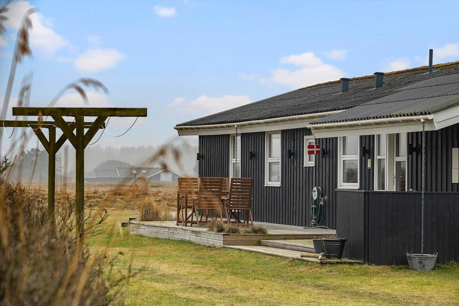 Black wooden house with terrace and garden. Wooden chairs are outside. Sky is blue with white clouds.