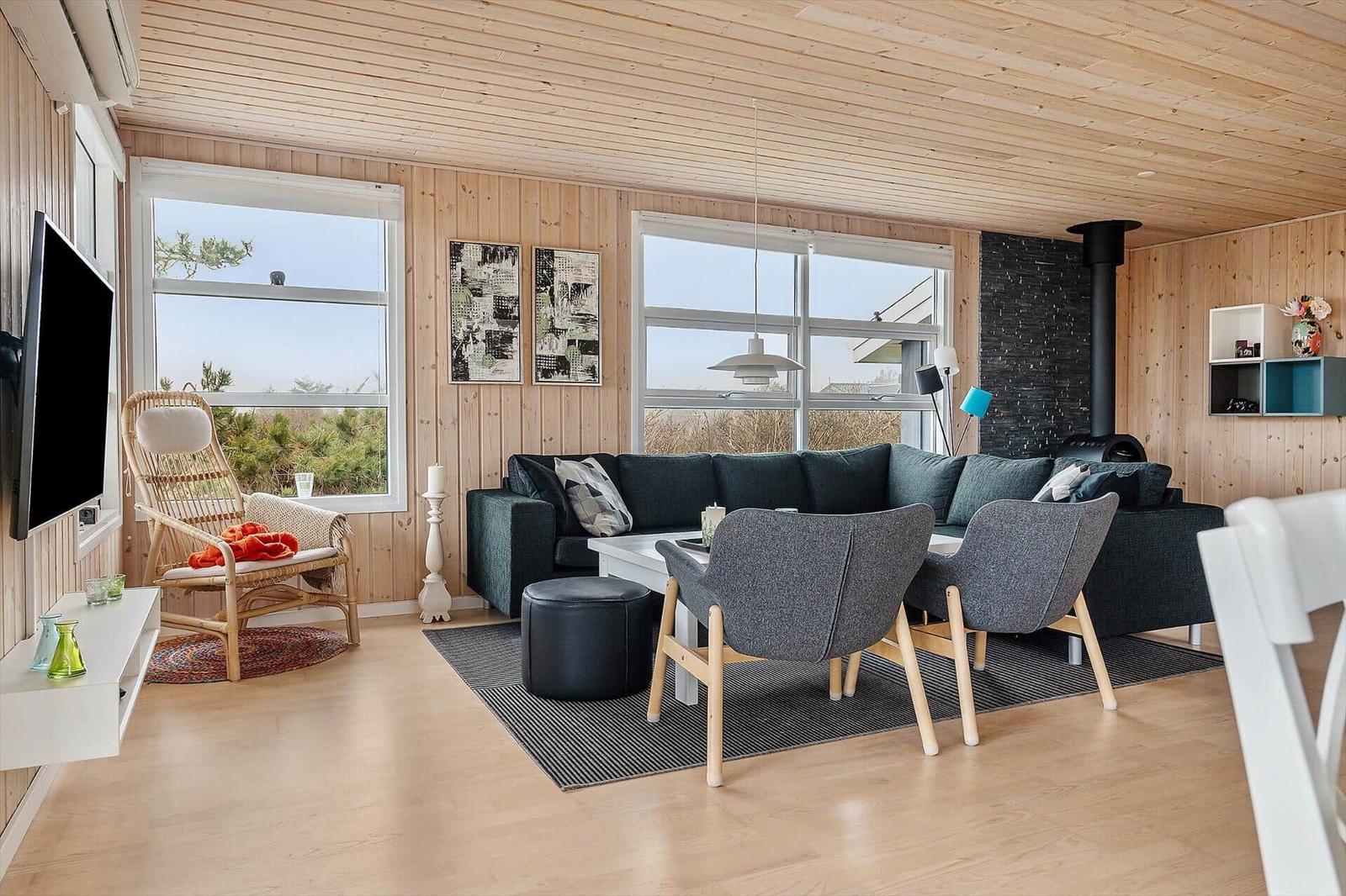 Living room with wooden walls, sofa, chairs, and TV. Large windows with view of green.