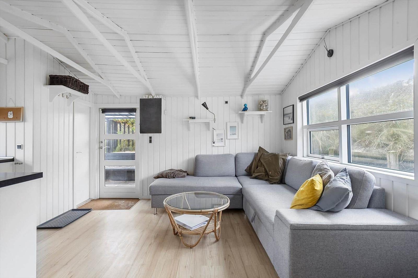 Wohnraum mit grauem Sofa, Glas-Tisch und großen Fenstern. Weiße Wände und Holzboden.