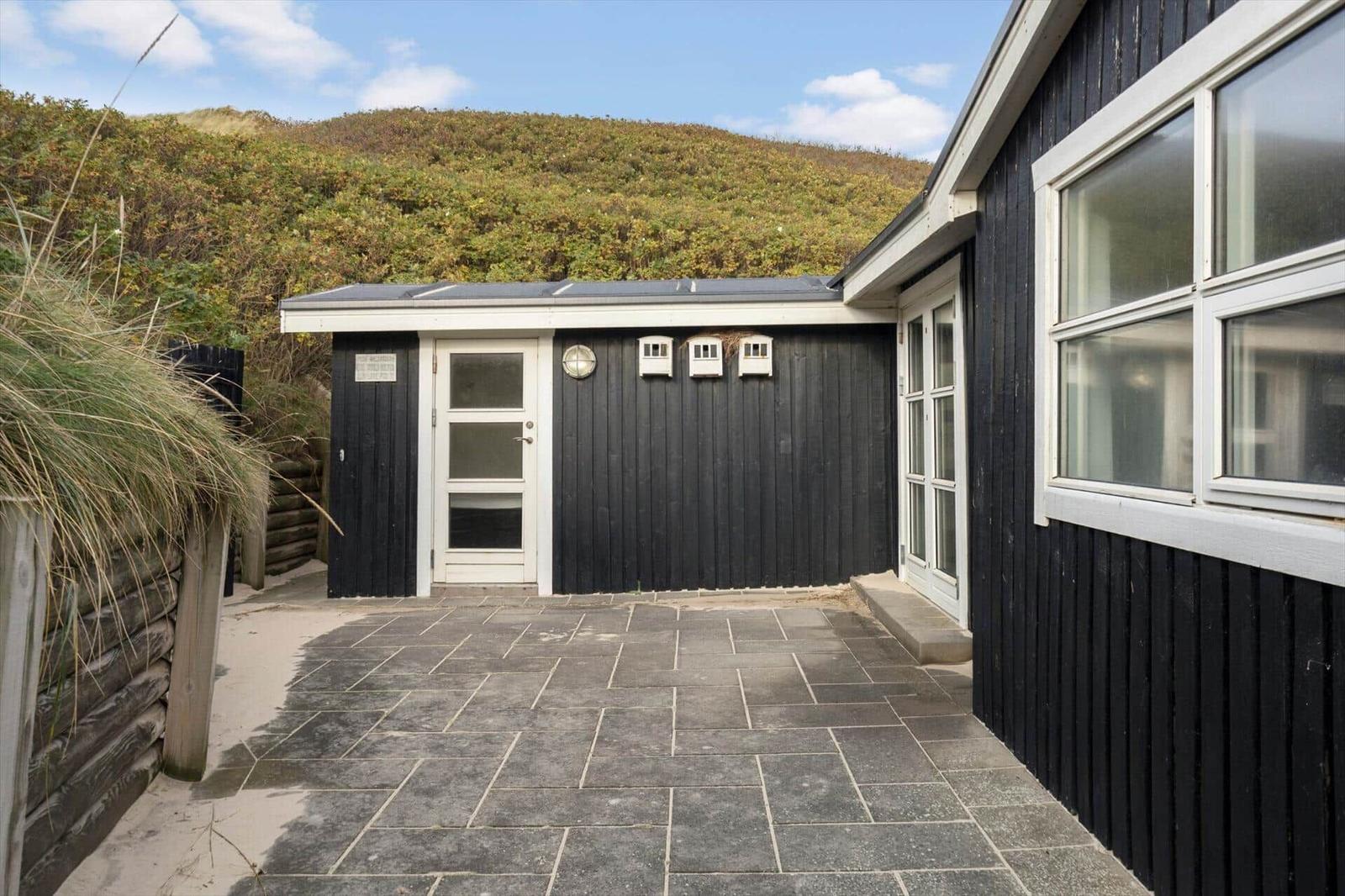 Schwarze Holzhütte mit weißer Tür und Fenster, gepflastertes Vorfeld, Hintergrund mit Dünenvegetation.