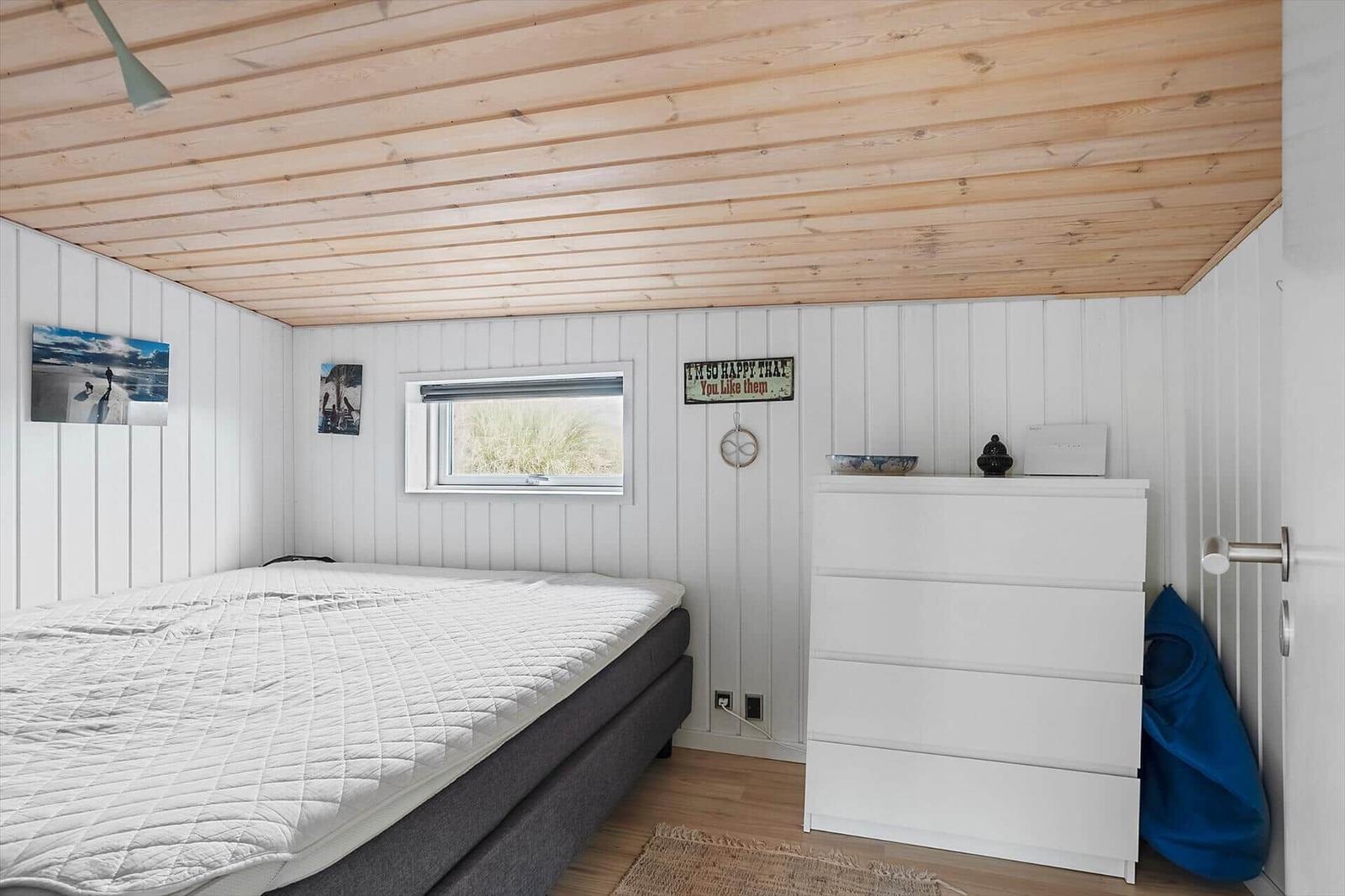 Schlafzimmer mit Bett, Kleiderschrank und Fenster. Holzdecke und weiße Wände.
