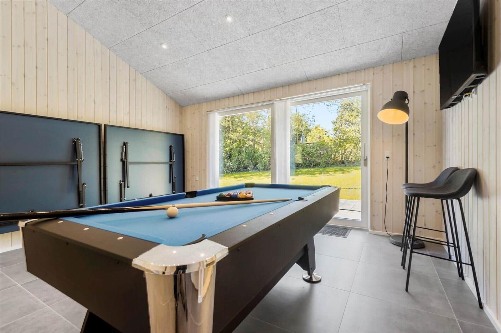 Pool table with blue felt, window to green outdoor area, two bar stools and wall-mounted TV.
