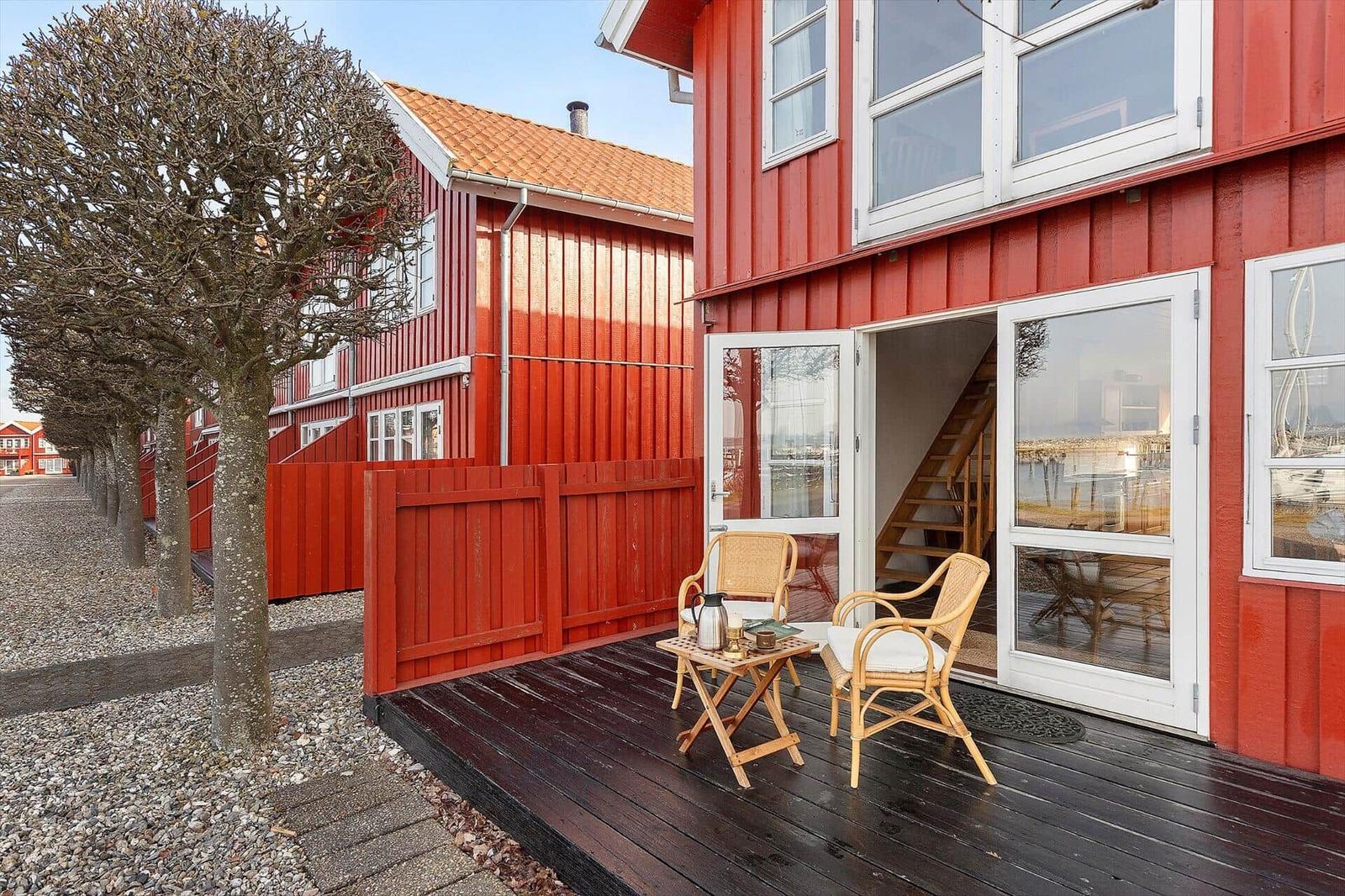 Red wooden house with terrace, two chairs, and table. Glass doors lead to interior.