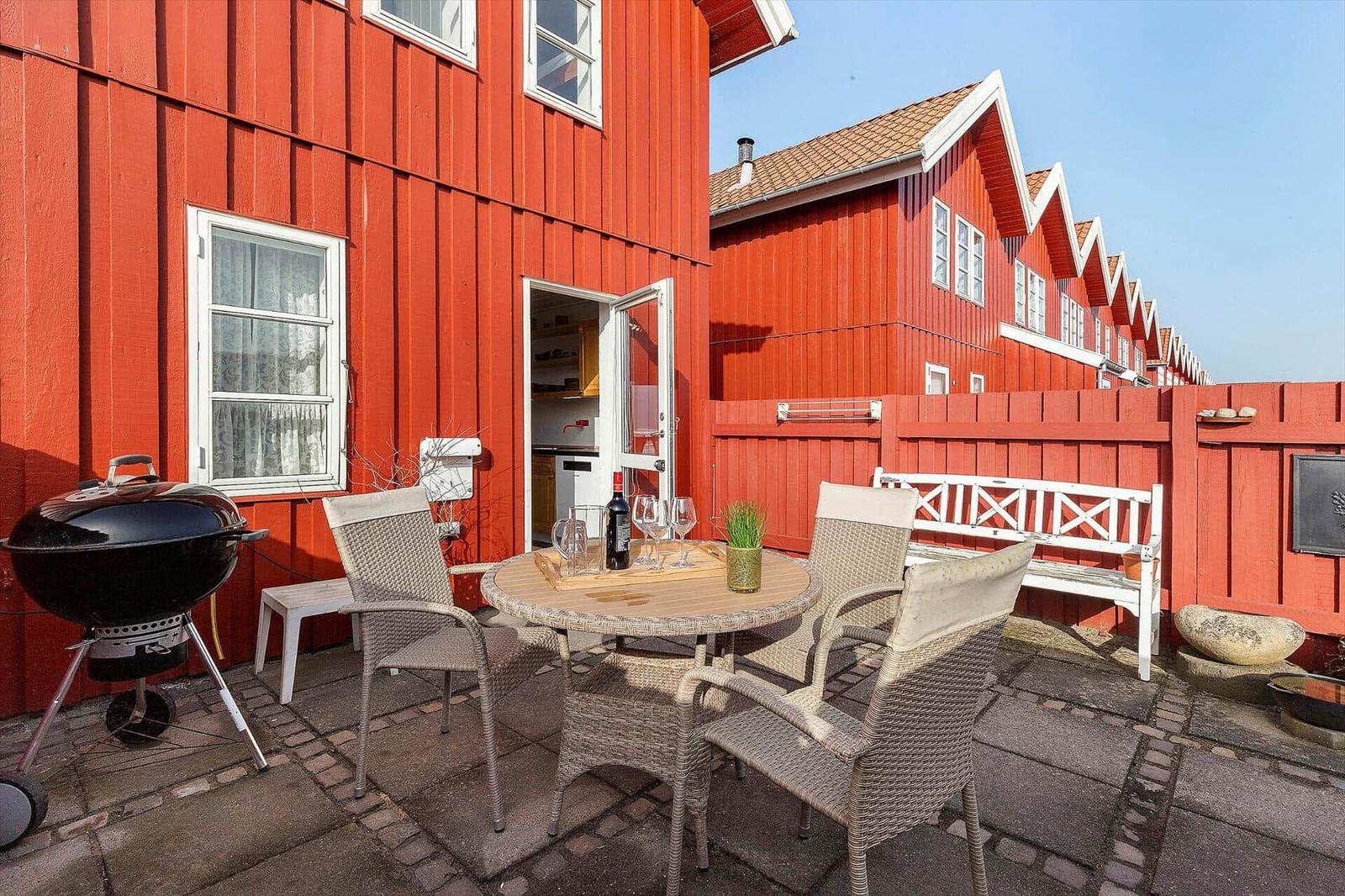 Red wooden house with terrace, grill, and seating. Backyard with white benches.