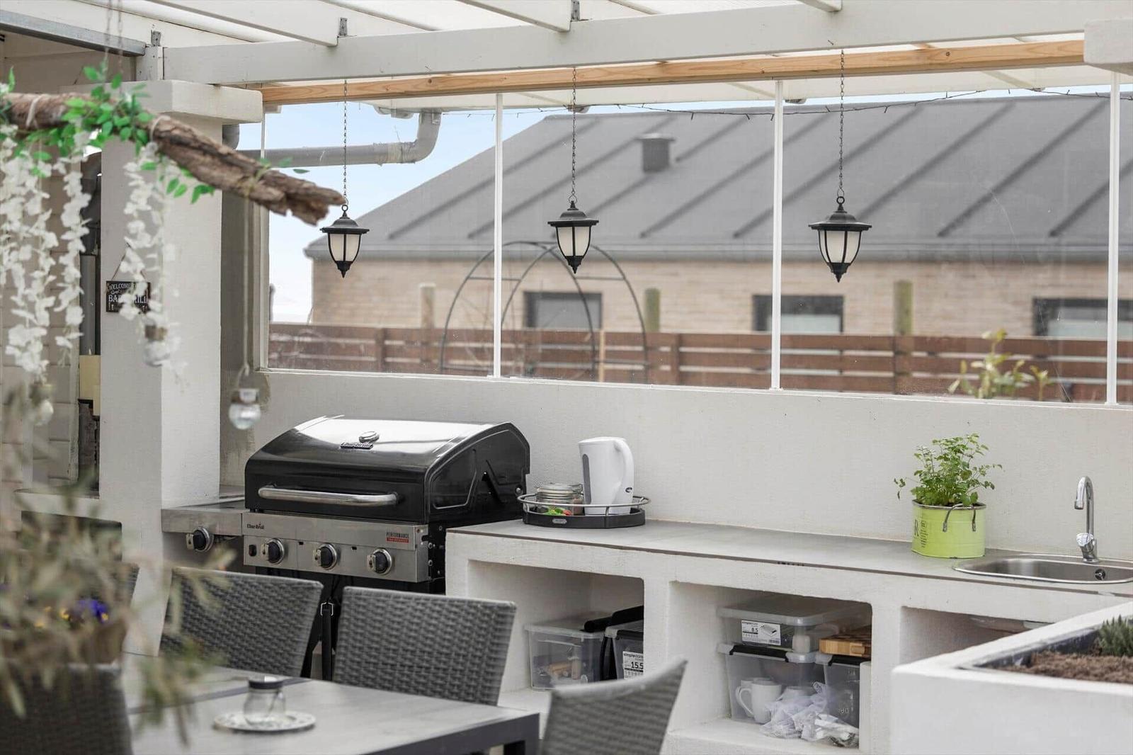 Terrasse mit Grill, Waschbecken und Hängelampen unter einer Markise.