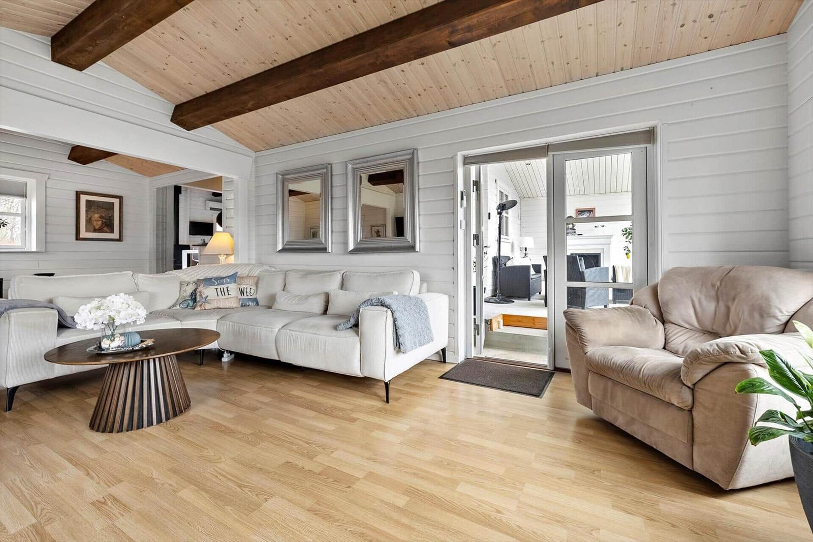 Wohnzimmer mit Sofa, Sessel und Holzboden. Deckenbalken und Glastür zur Terrasse.