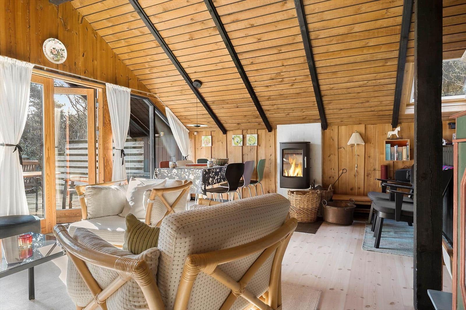 Wohn- und Essbereich mit Kamin, Holzwand und großem Fenster. Rattanmöbel und warmes Licht.