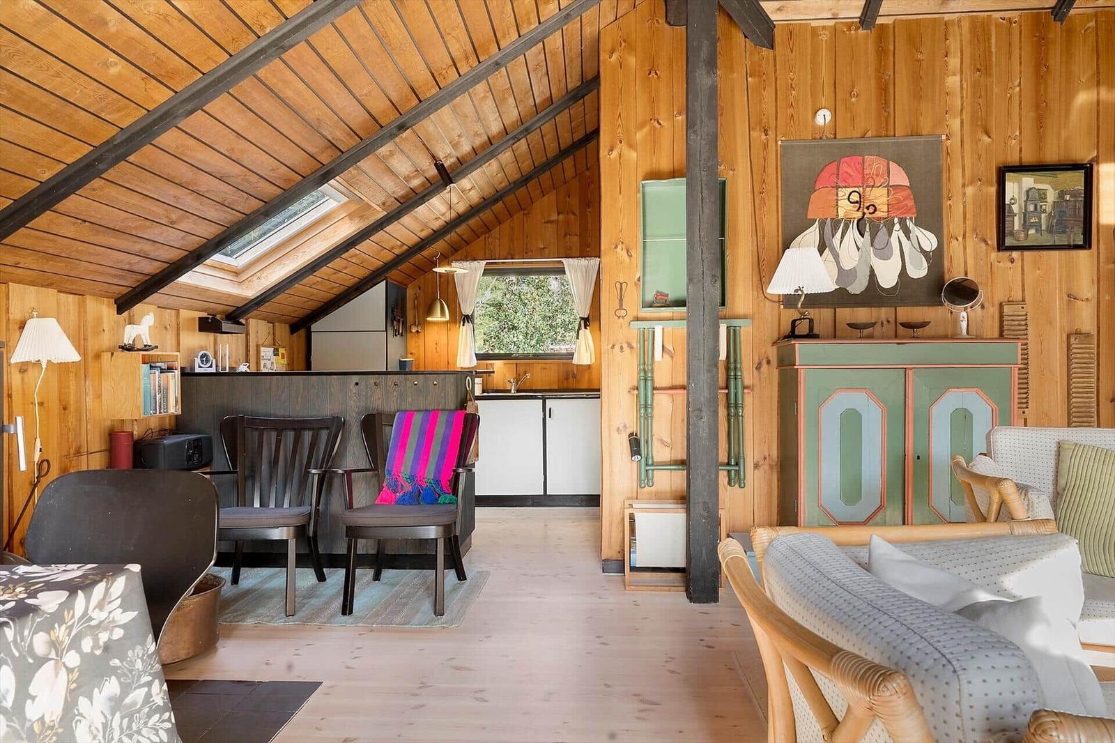 Wohnbereich mit Holzwand, Sofa, Stühlen und Küchenbereich unter Dachschrägen.