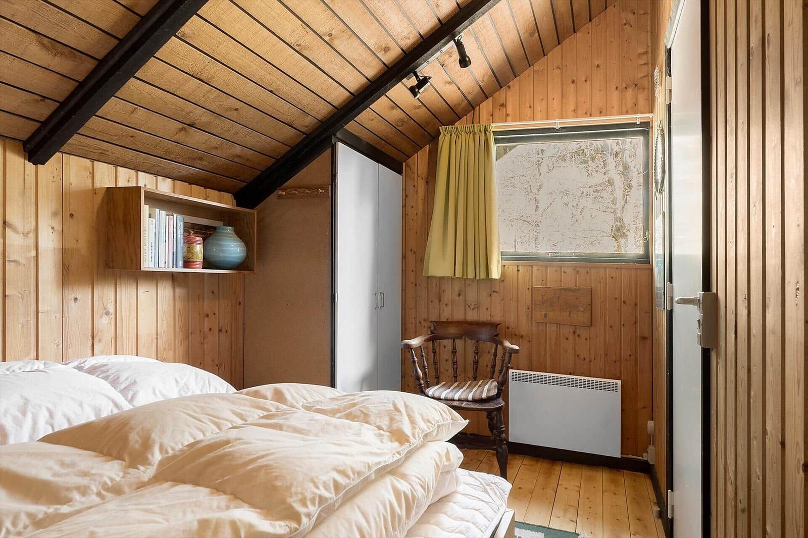 Schlafzimmer mit Holzwand, Bett, Stuhl und Fenster mit Vorhang.