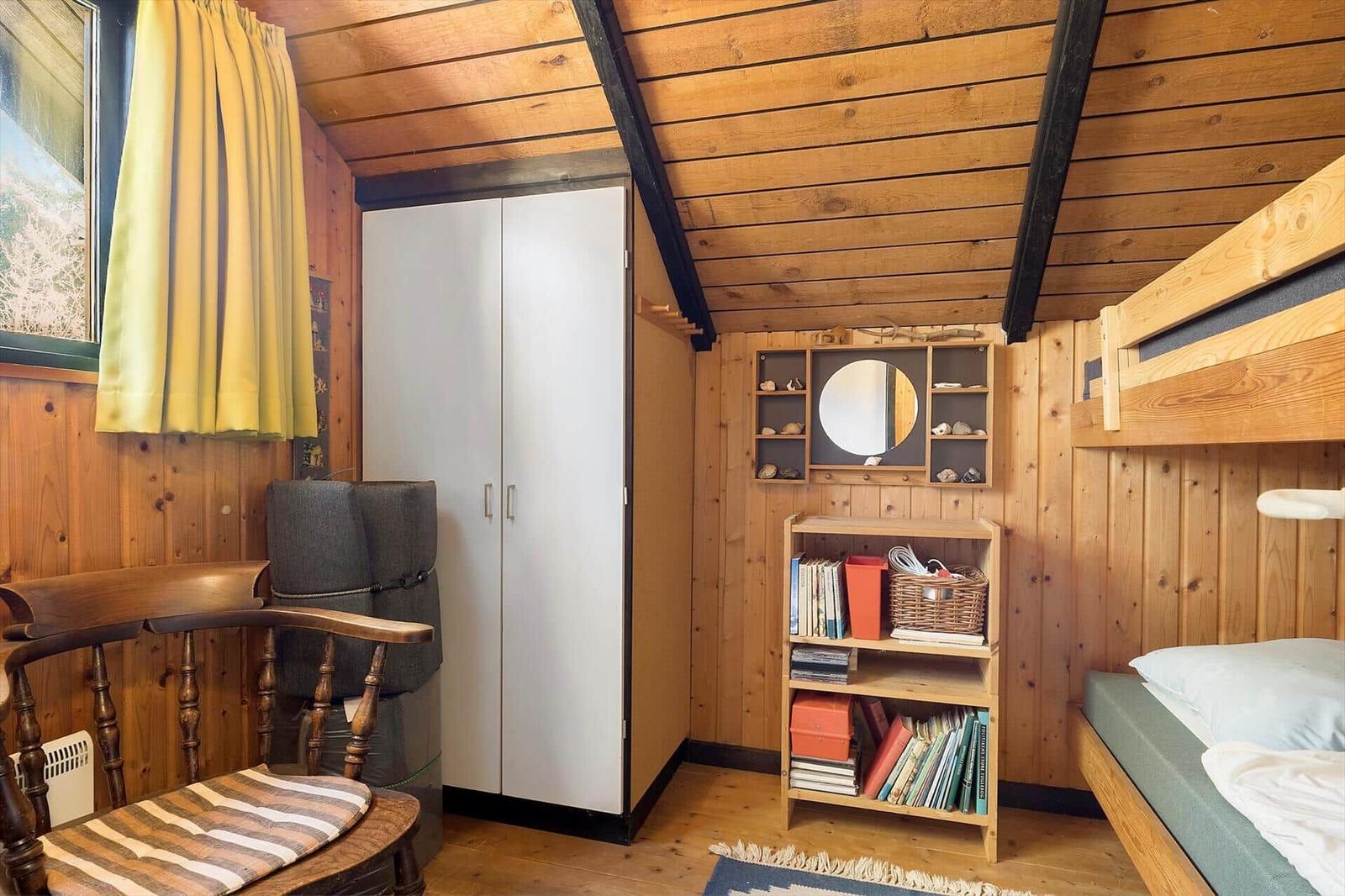 Zimmer mit Holzwand, Doppelbett, Schrank, Bücherregal und Fenster mit gelben Vorhängen.