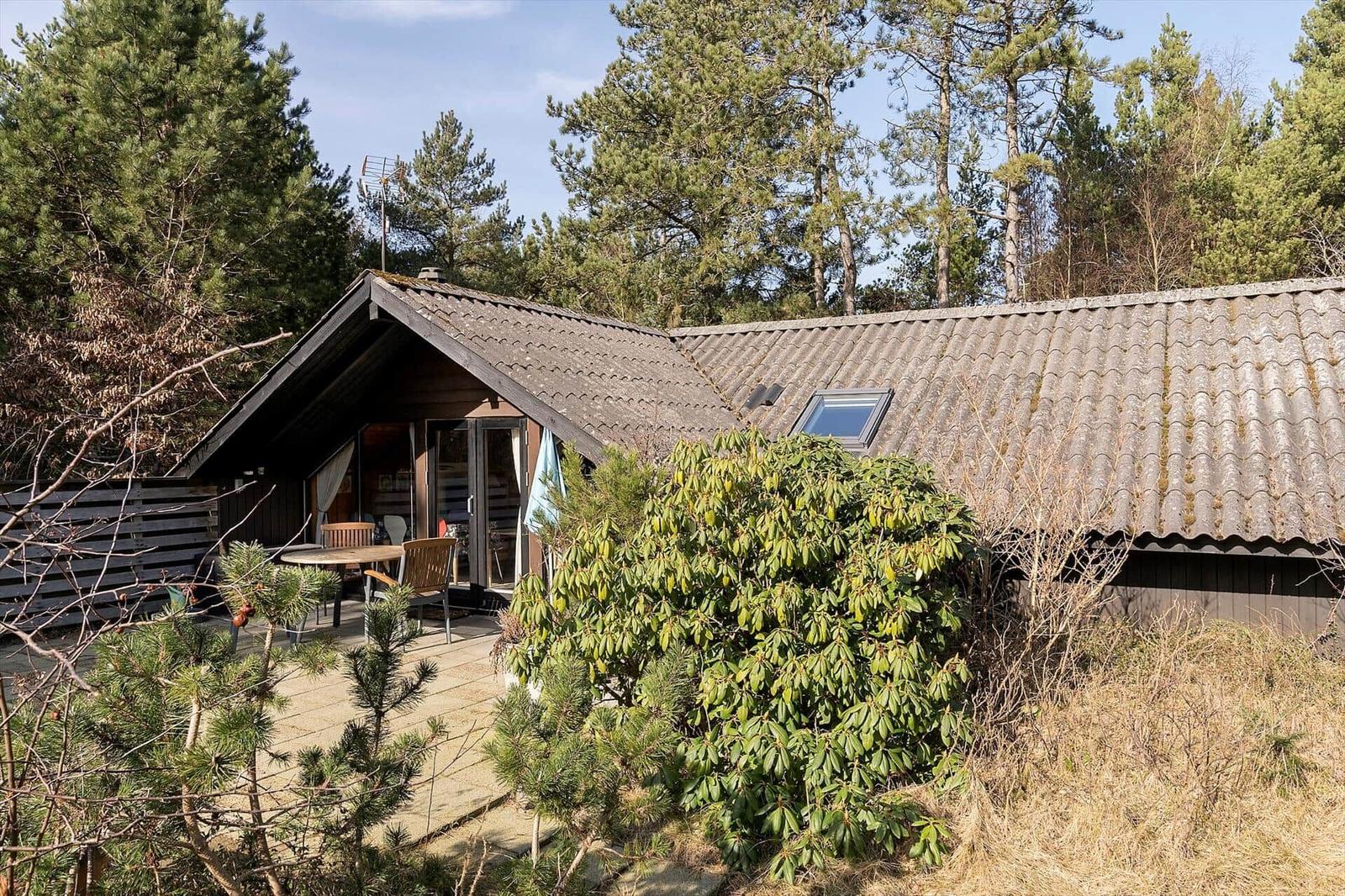 Holzhaus mit Terrasse, Garten und Dachfenster. Umgeben von Bäumen und Sträuchern.