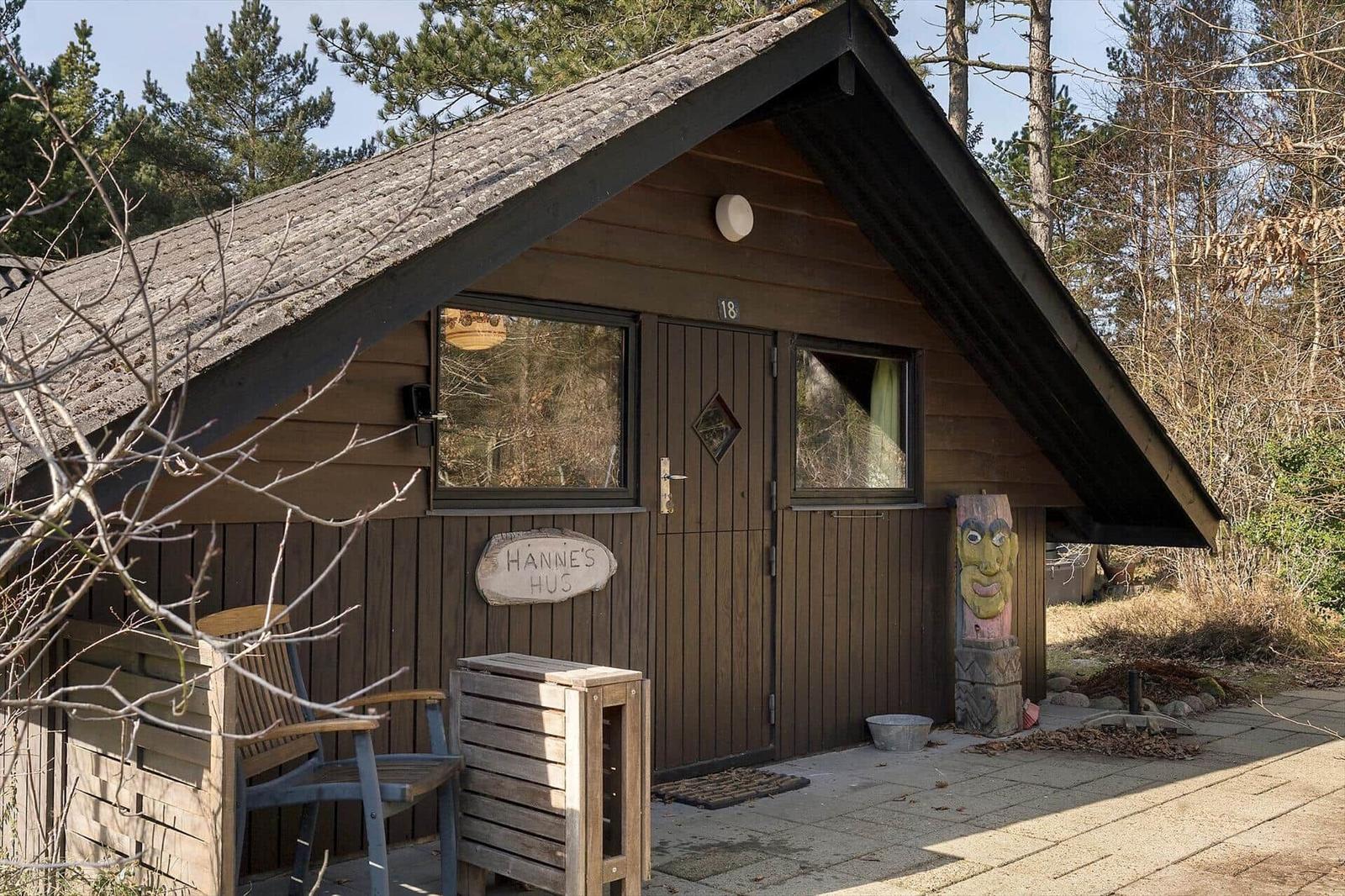 Hütte mit Holzfassade, Tür und Fenstern. Vor dem Haus steht ein Stuhl und eine Skulptur.