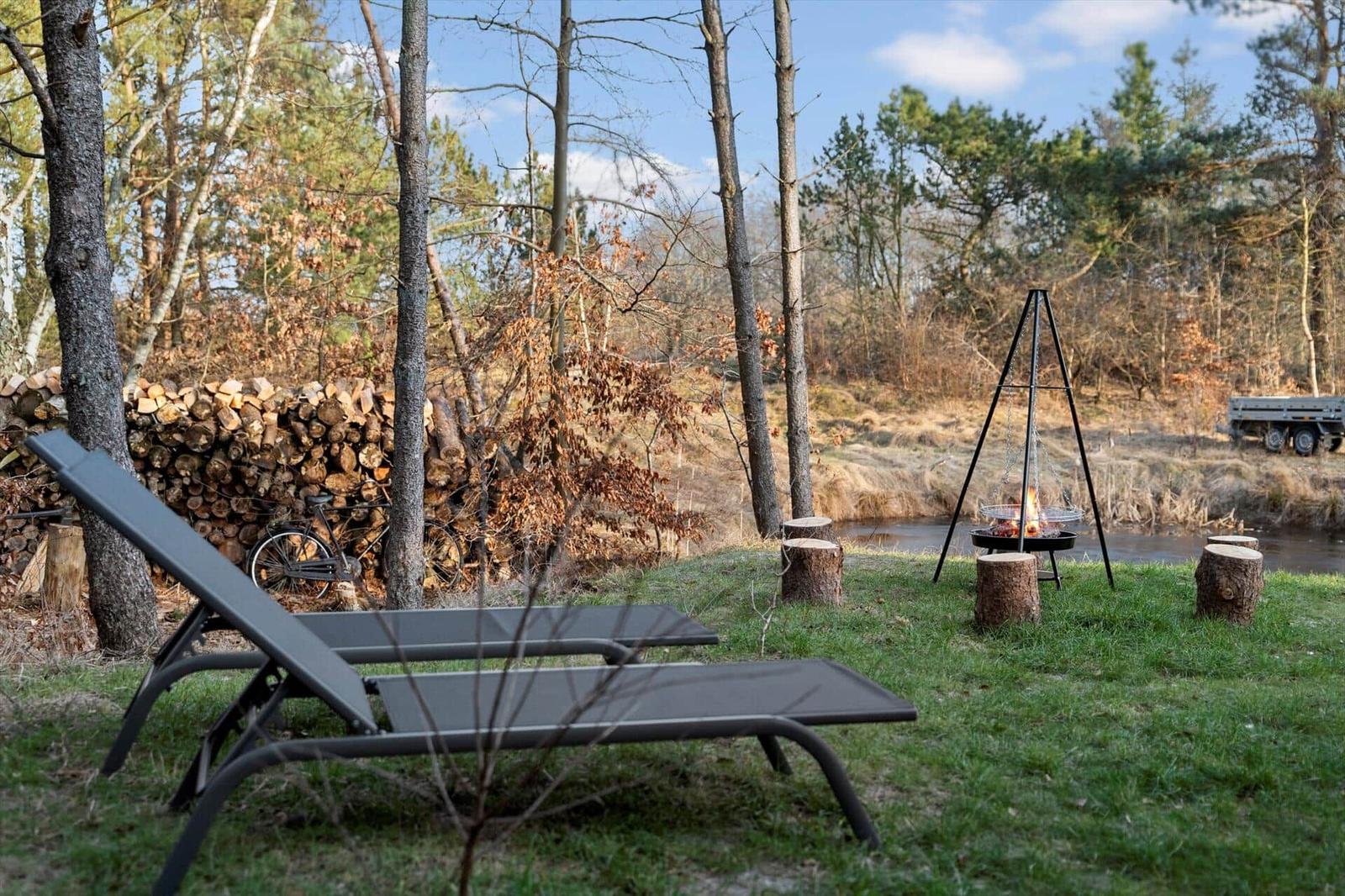 Two lounge chairs next to a grill in the woods. Background: woodpile and river.