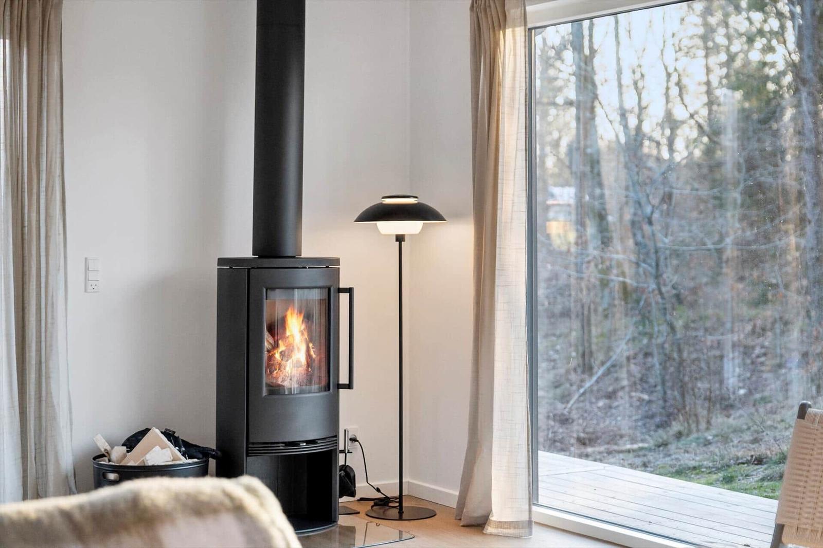 Wood-burning stove, large window with forest view, floor lamp, and curtains.