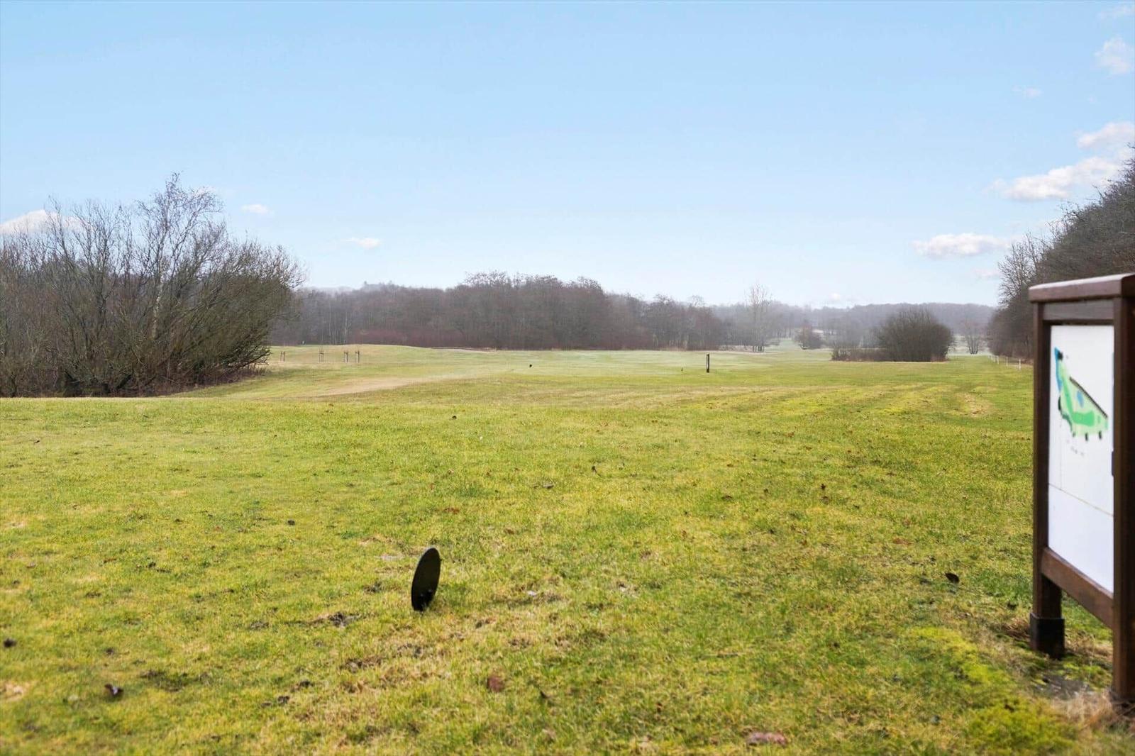 Golfplatz mit grünem Rasen und Bäumen im Hintergrund. Informationstafel rechts.