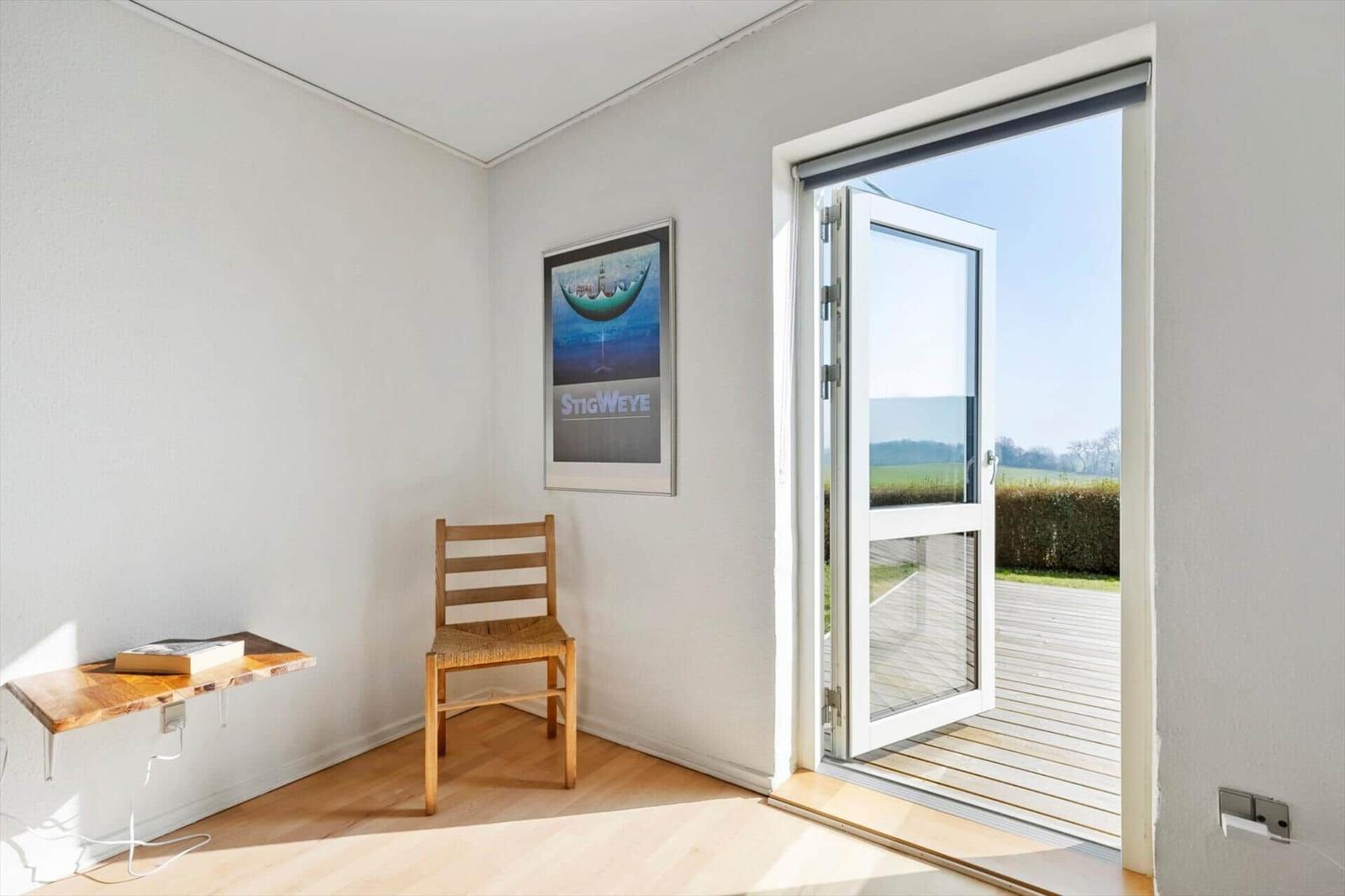 Room with chair, table, and poster on wall. Through glass door, terrace and landscape are visible.