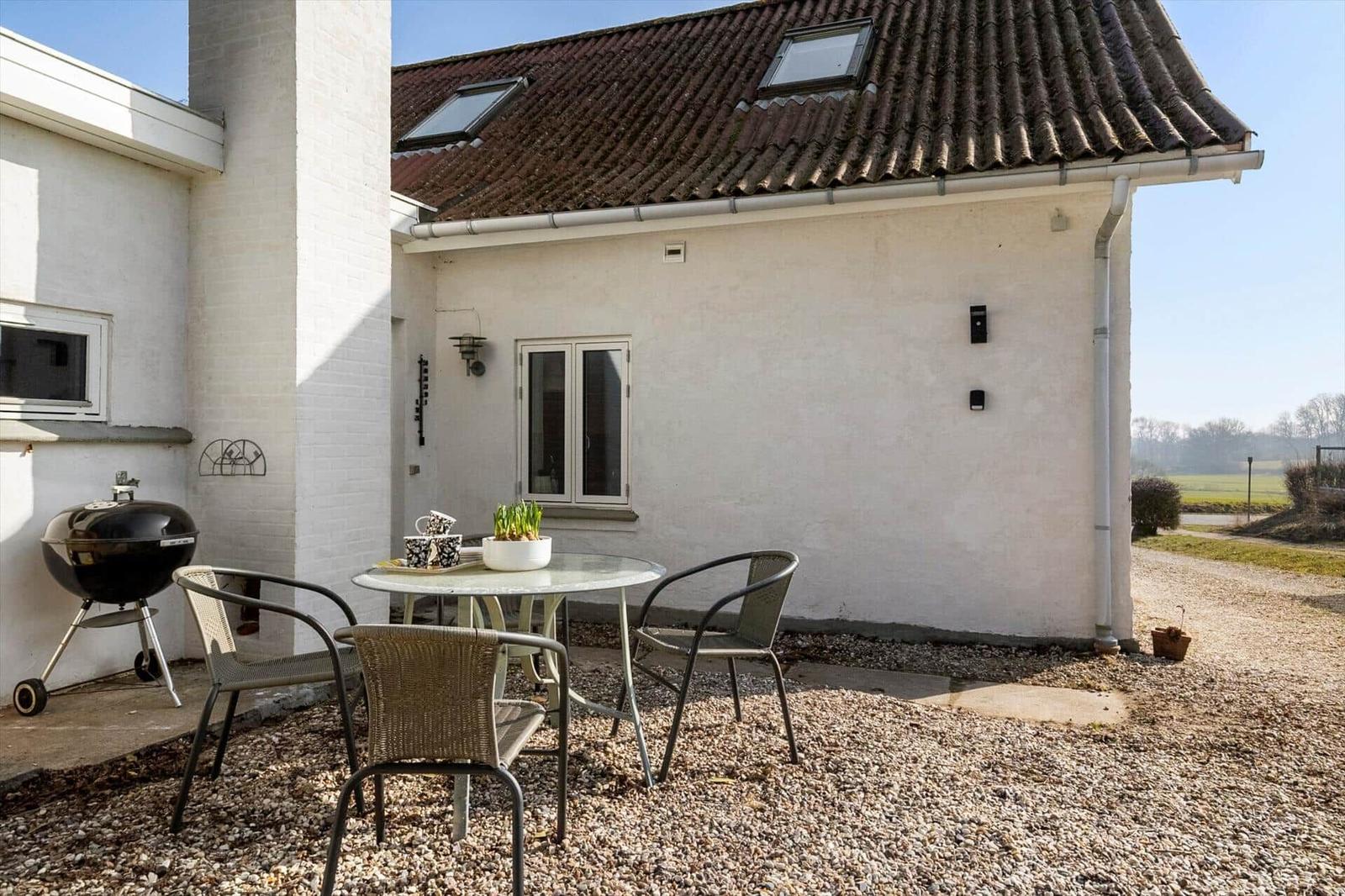 Outdoor area with table, chairs, and grill beside white wall and roof.