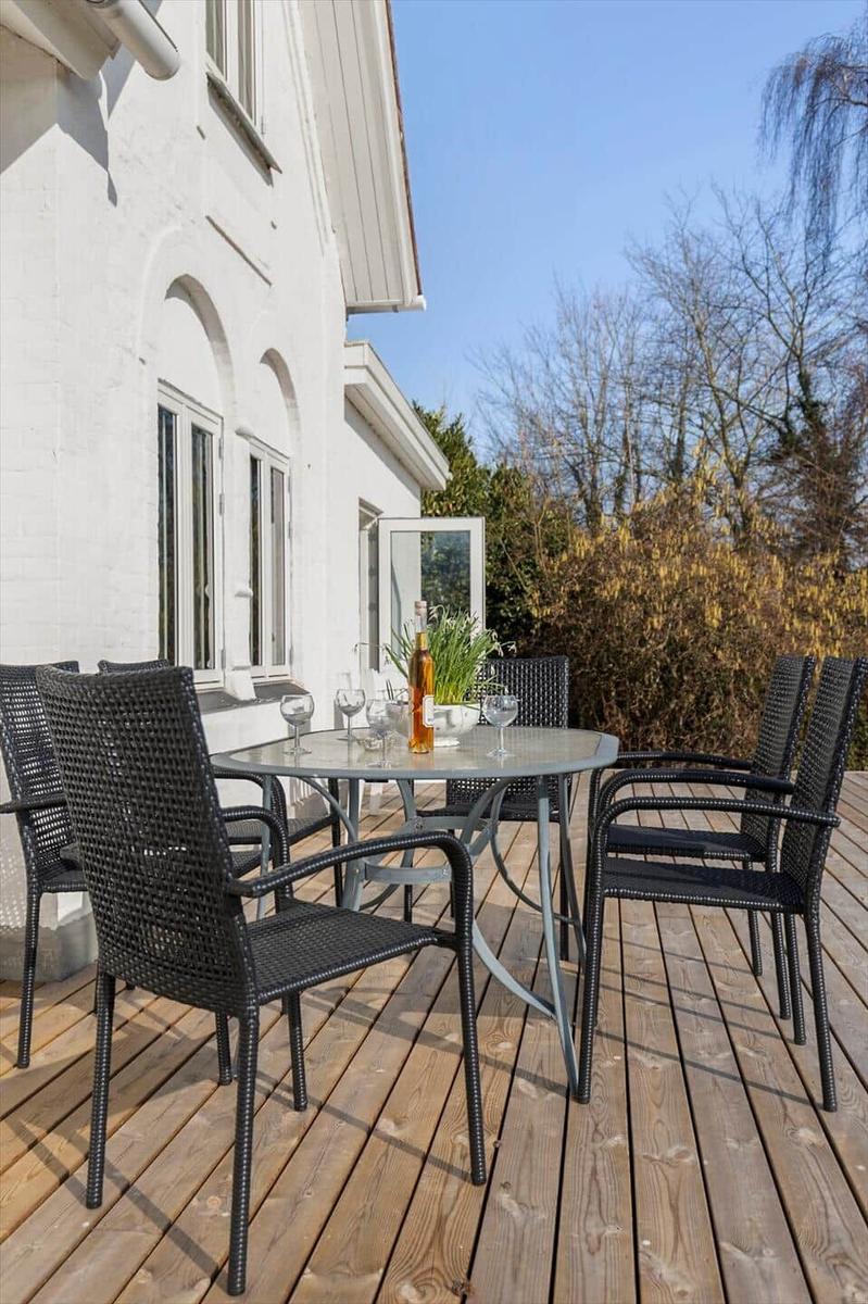 Deck with table and chairs, wooden floor, white house wall, trees in background.