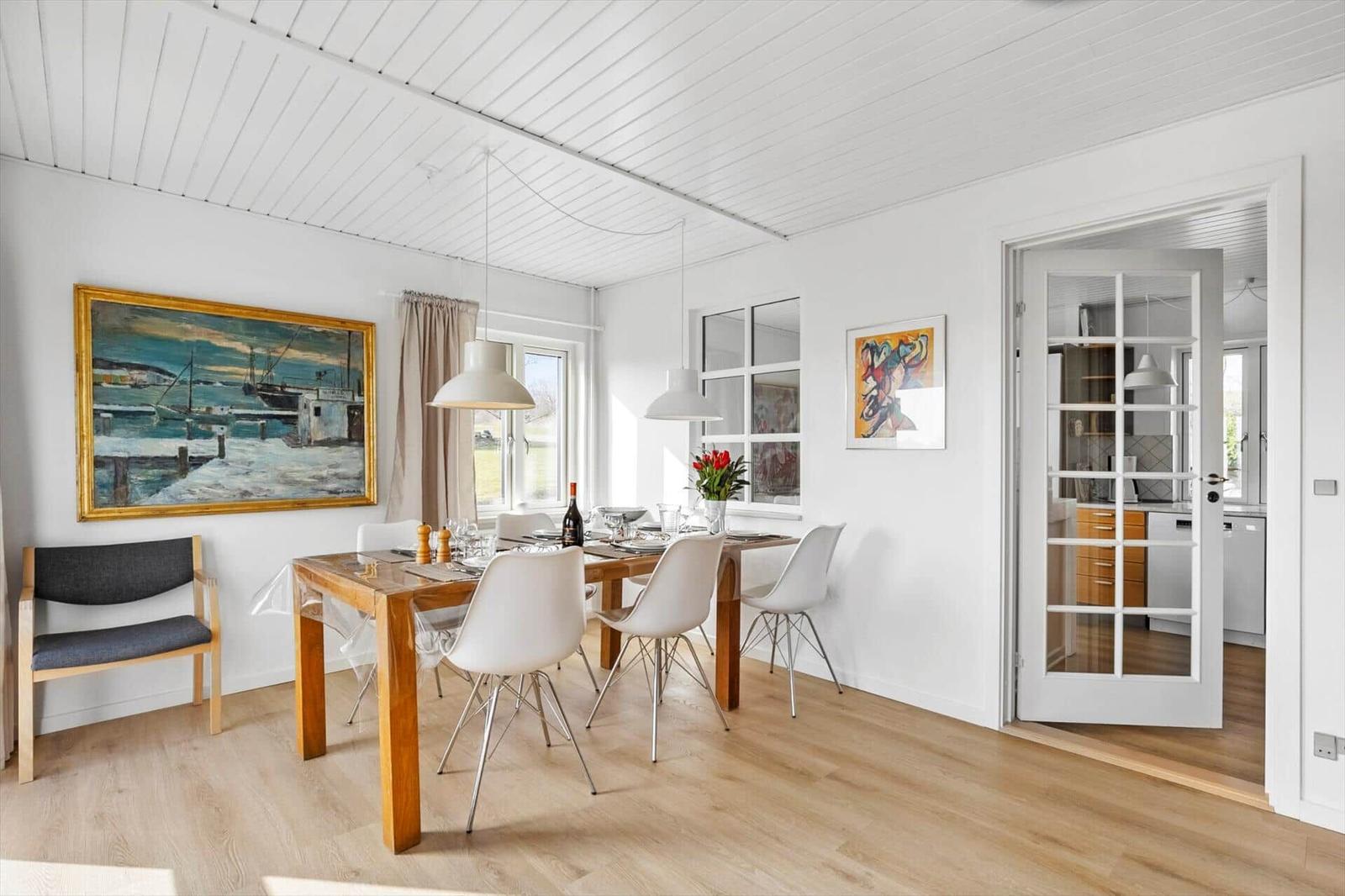 Dining area with wooden table, white chairs, and paintings on walls. Passage to kitchen.