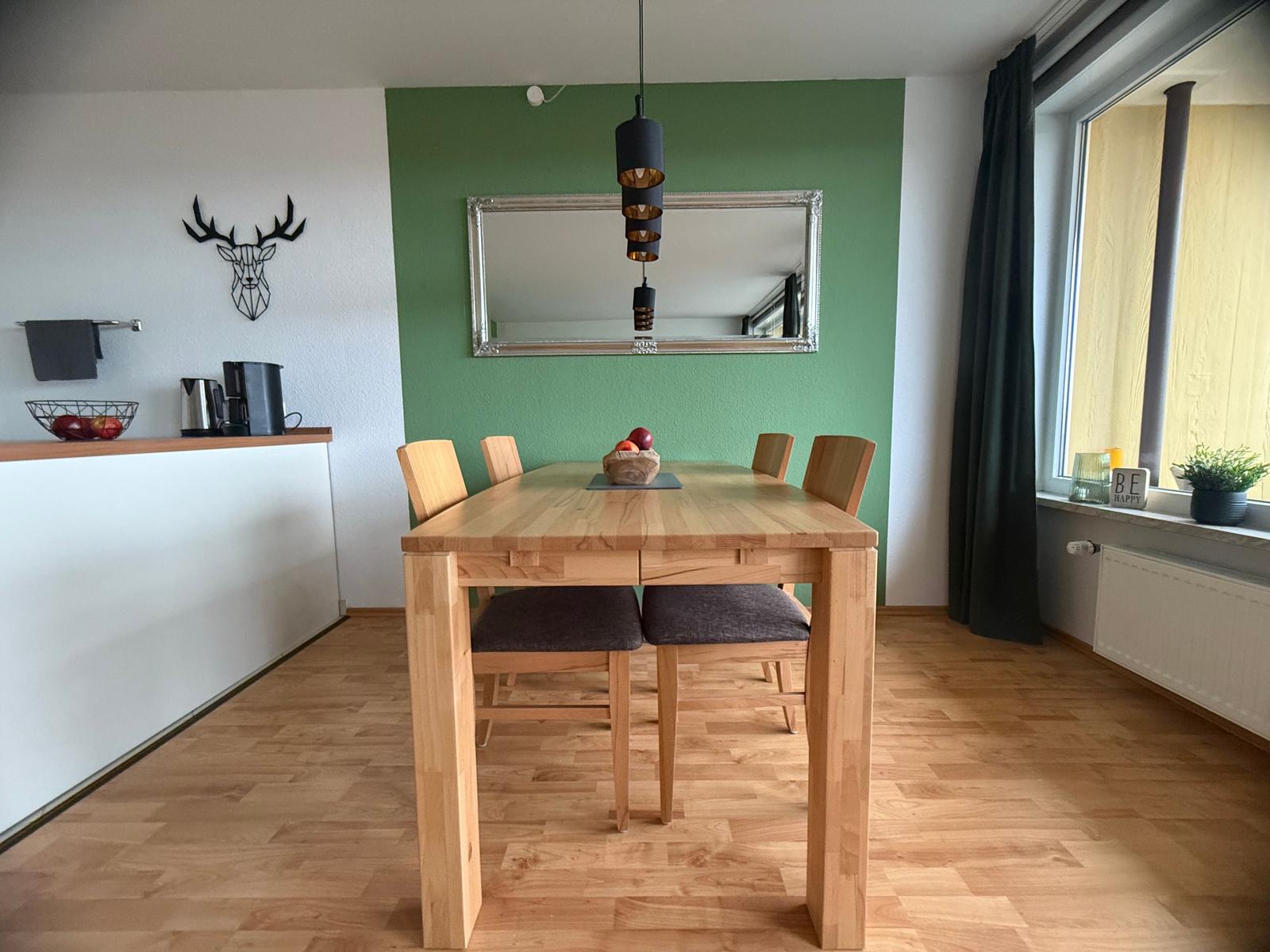 Esszimmer mit Holztisch, Stühlen und grünem Wandbereich. Fenster mit Vorhang und Pflanze.