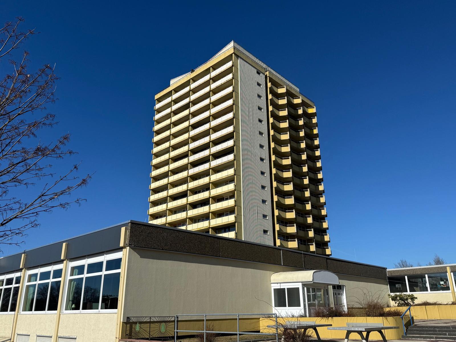 Hochhaus mit Balkonen und kleinem Nebengebäude unter blauem Himmel.