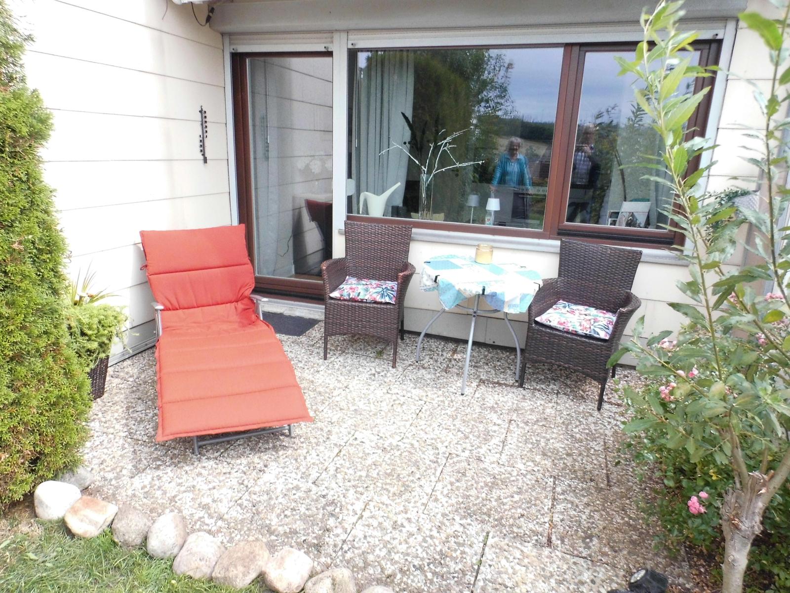 Terrasse mit Liegestuhl, zwei Stühlen und Tisch. Grünflächen und Steine umgeben den Platz.