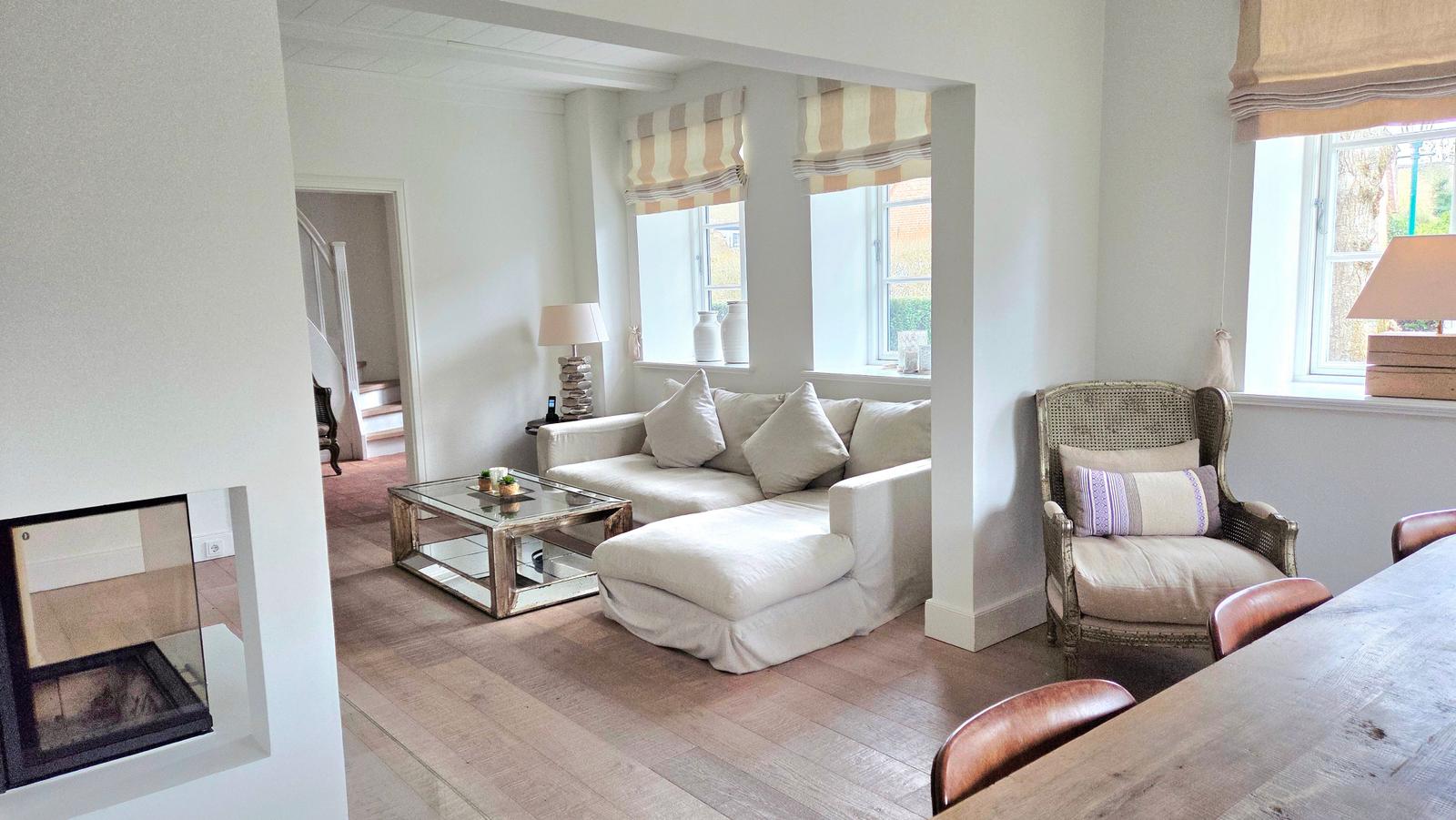 Wohnbereich mit Sofa, Sessel und Esstisch. Durchgang zur Treppe und Fenster mit Rollläden.