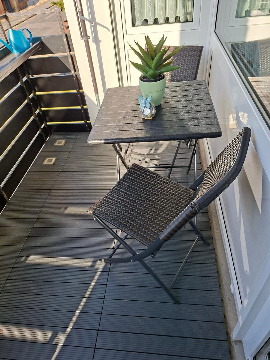 Terrasse mit Tisch, zwei Stühlen und Pflanze. Holzdielen und Fenster im Hintergrund.