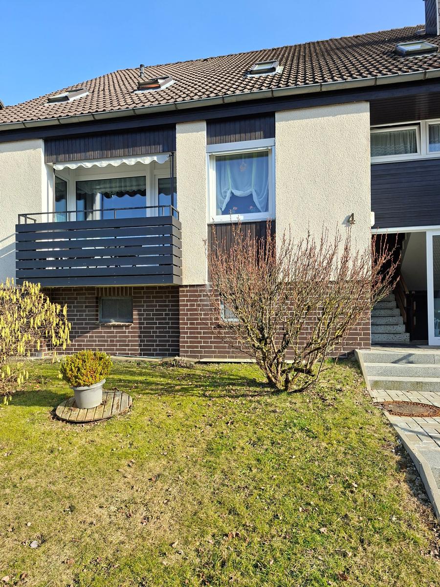 Haus mit Balkon, Garten und Eingangstreppe. Fenster mit Vorhängen.